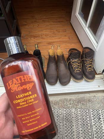 reviewer holding up the bottle of leather conditioner in front of several pairs of leather boots