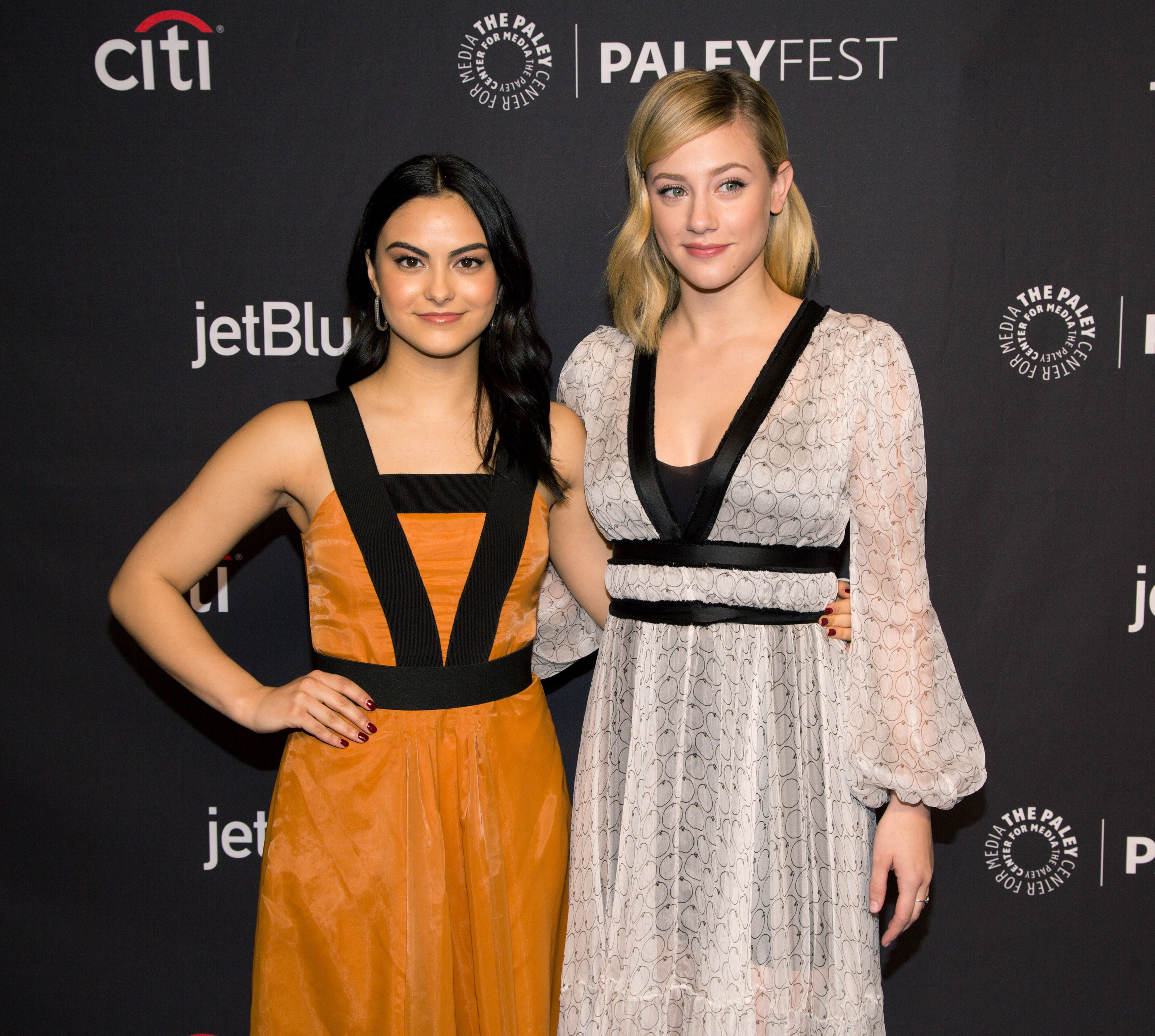 Camila Mendes in an orange sleeveless dress with black straps forming a V shape from shoulder to waist. Her dark hair is down and she smiles with her arm around Lili Reinhart, who wears her hair down and a white chiffon-like dress with balloon sleeves.