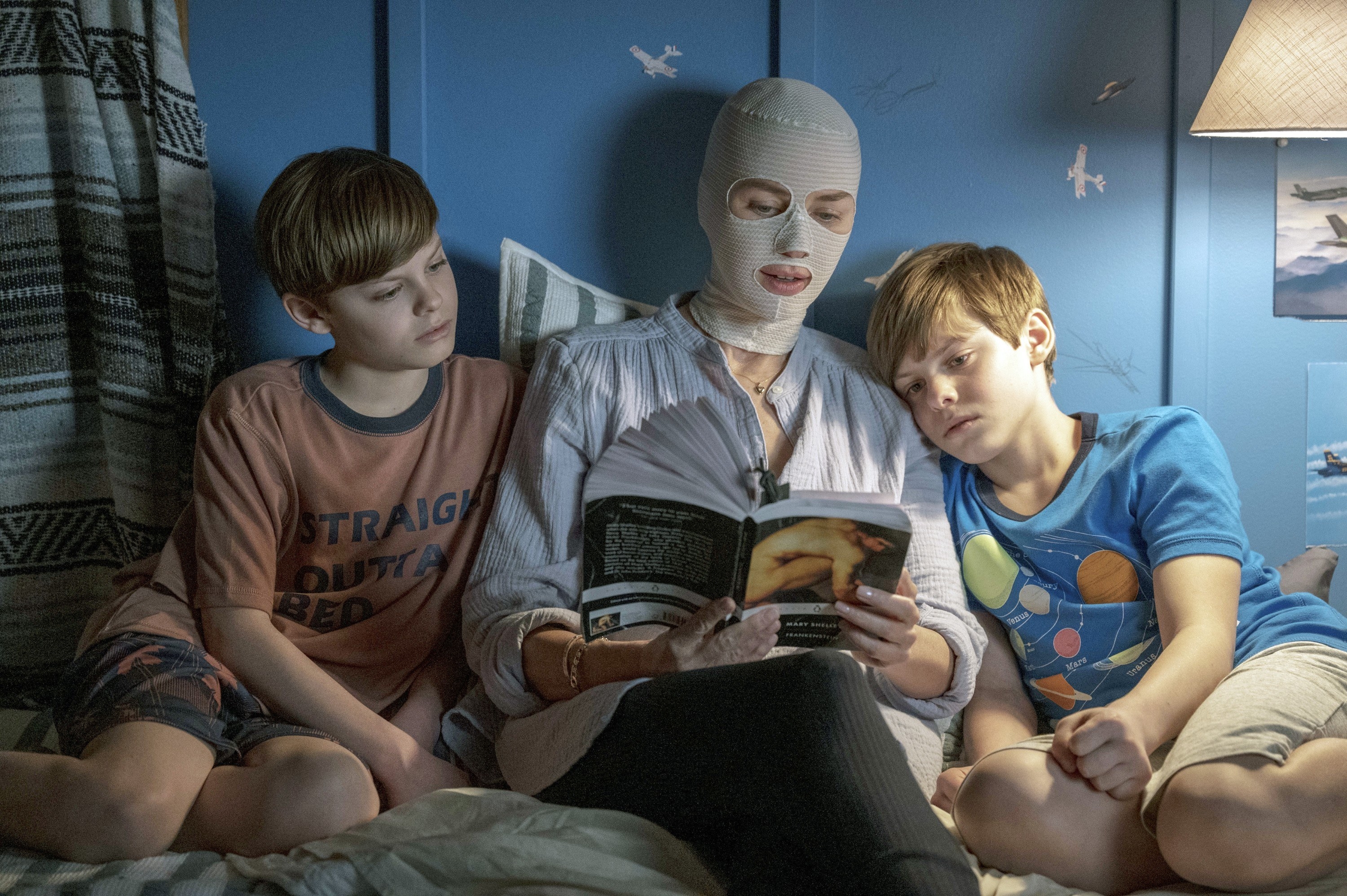 A woman in a strange white mask sits in bed reading a book to two small boys