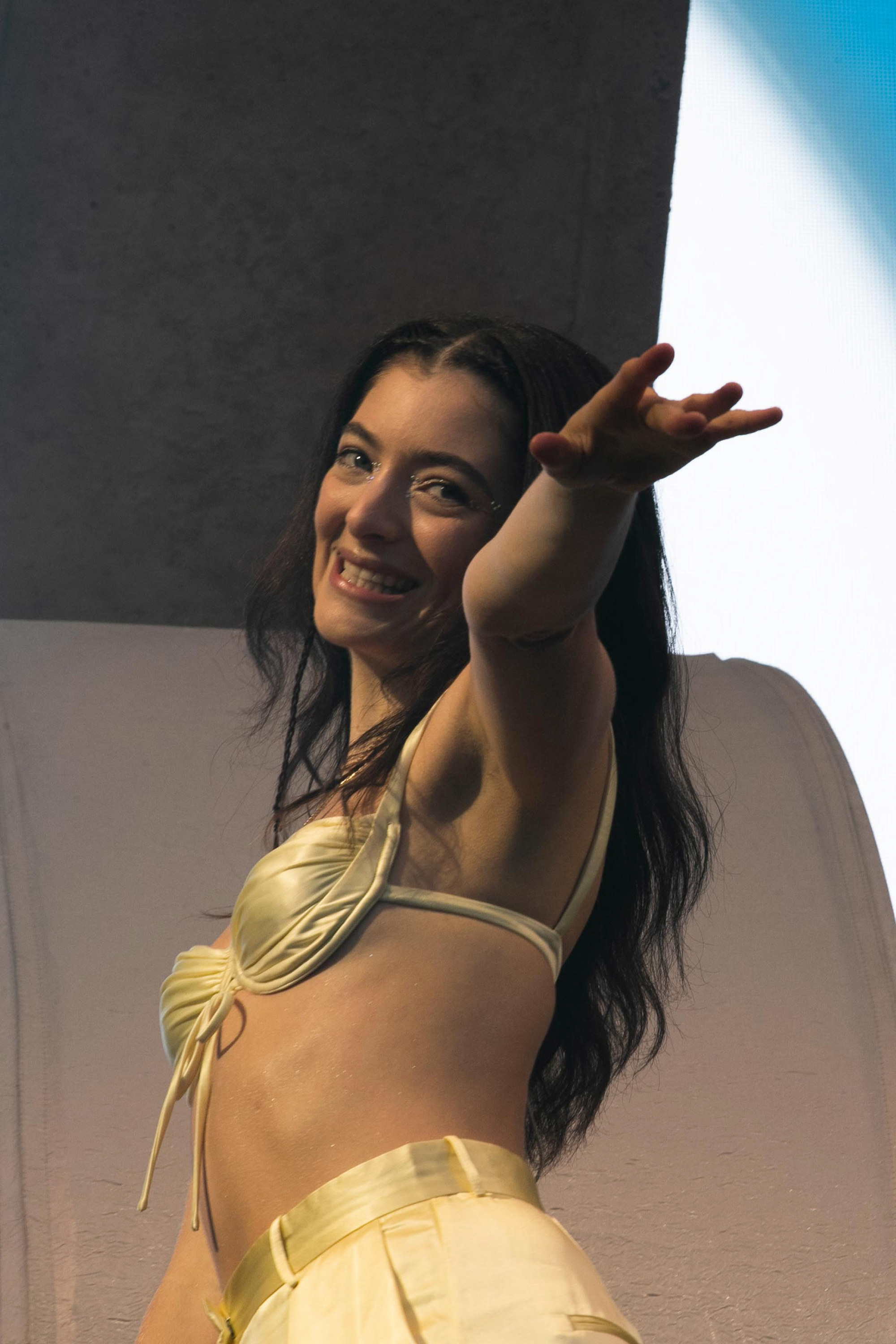 Image of Lorde in a lemon yellow bikini top, grinning and waving from on stage