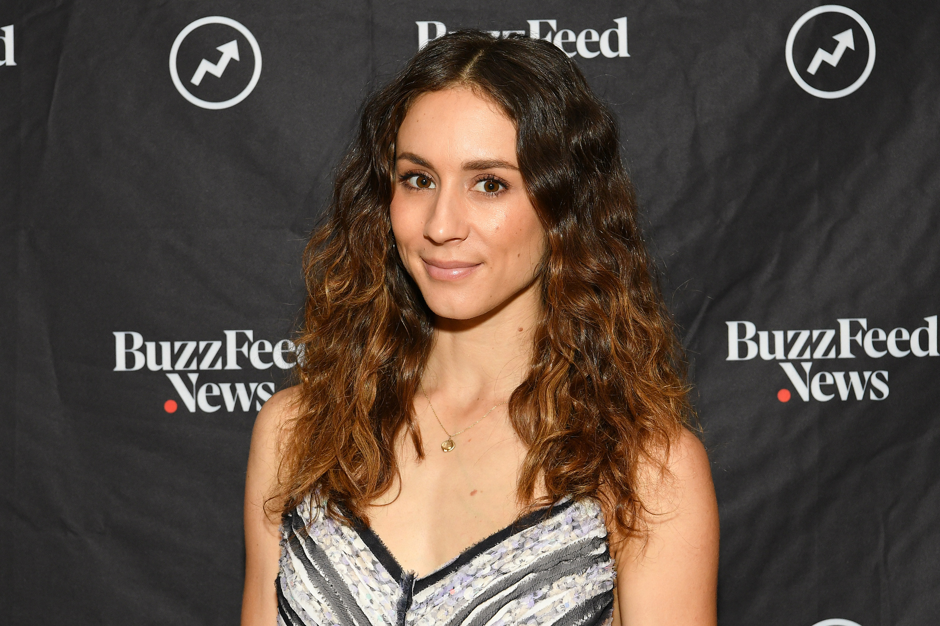 Head and shoulders shot of Troian with wavy dark ombre hair, smiling. She wears a grey and black v neck top