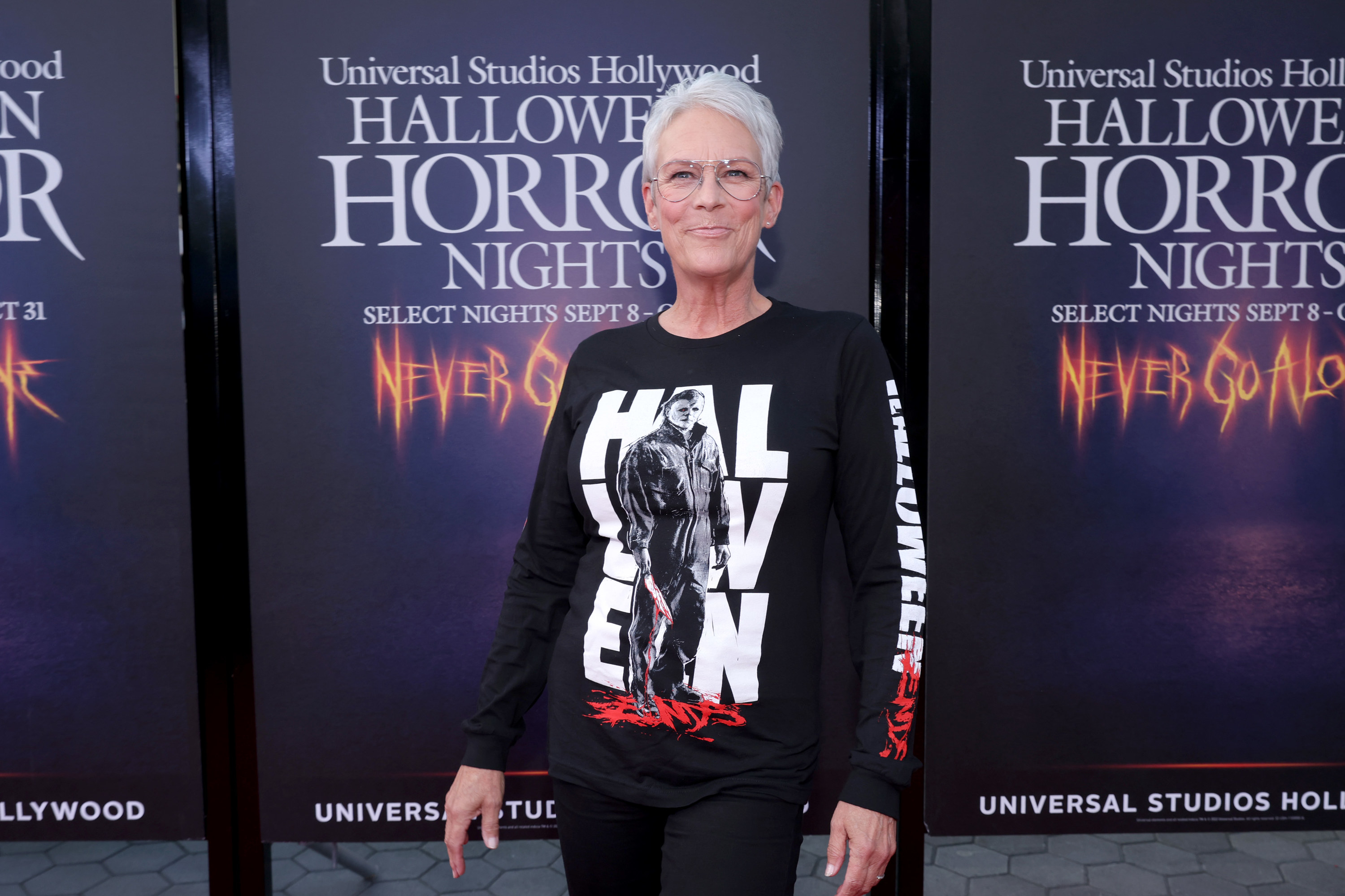 Jamie Lee Curtis wearing a &quot;Halloween&quot; long sleeved t-shirt with Michael Myers on it, smiling in front of a poster for Universal Studios Hollywood Halloween Horror Nights