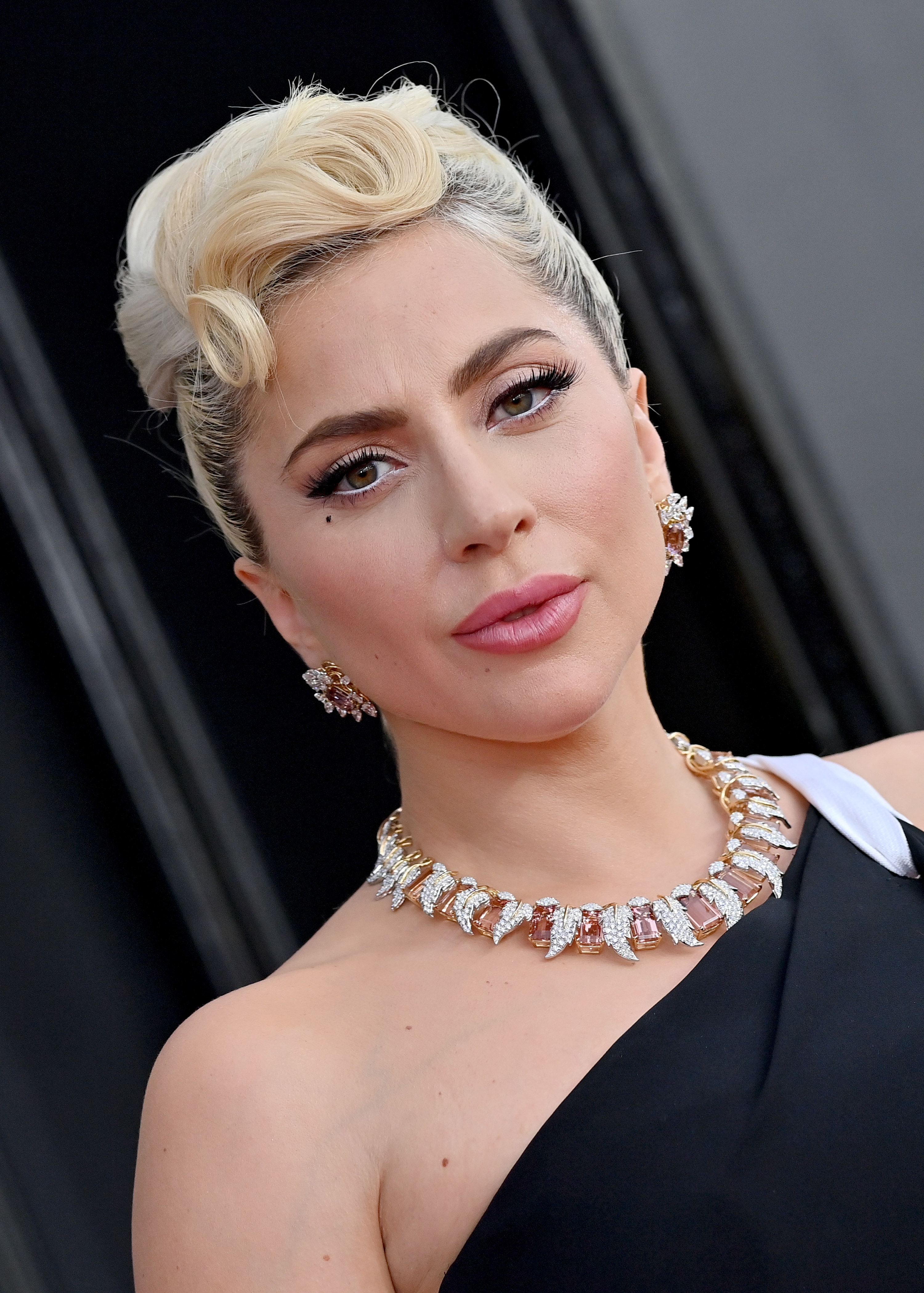 Head and shoulders shot of Lady Gaga smiling, her blonde hair swept up in a tight bun and her eye make up accentuating her eyes with white liner and black lashes. She wear chunky jewelled earrings and a matching necklace.