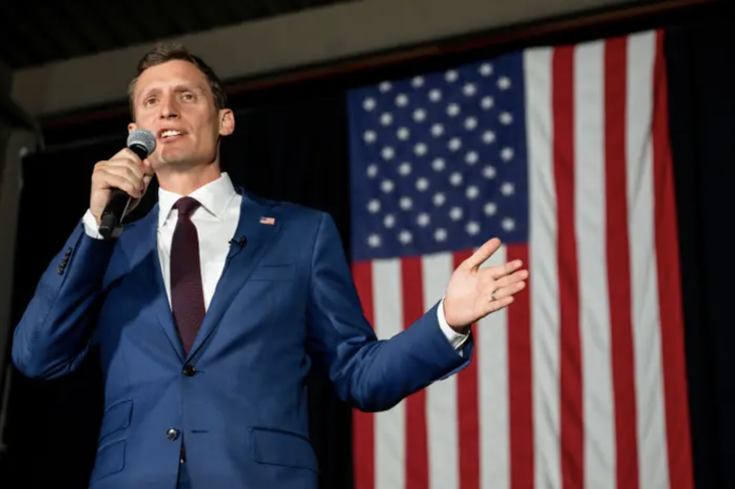 Seth Masters wears a blue suit and gestures while speaking into a microphone in front of an American flag