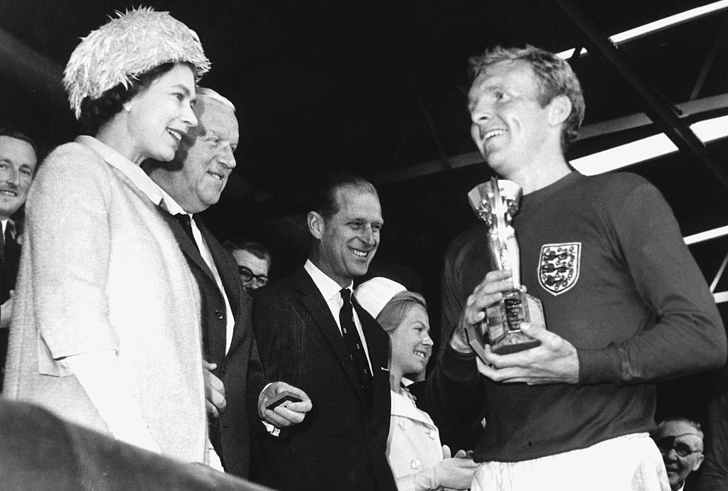 the queen handing over a trophy to bobby moore whilst standing in the stands of a football stadium