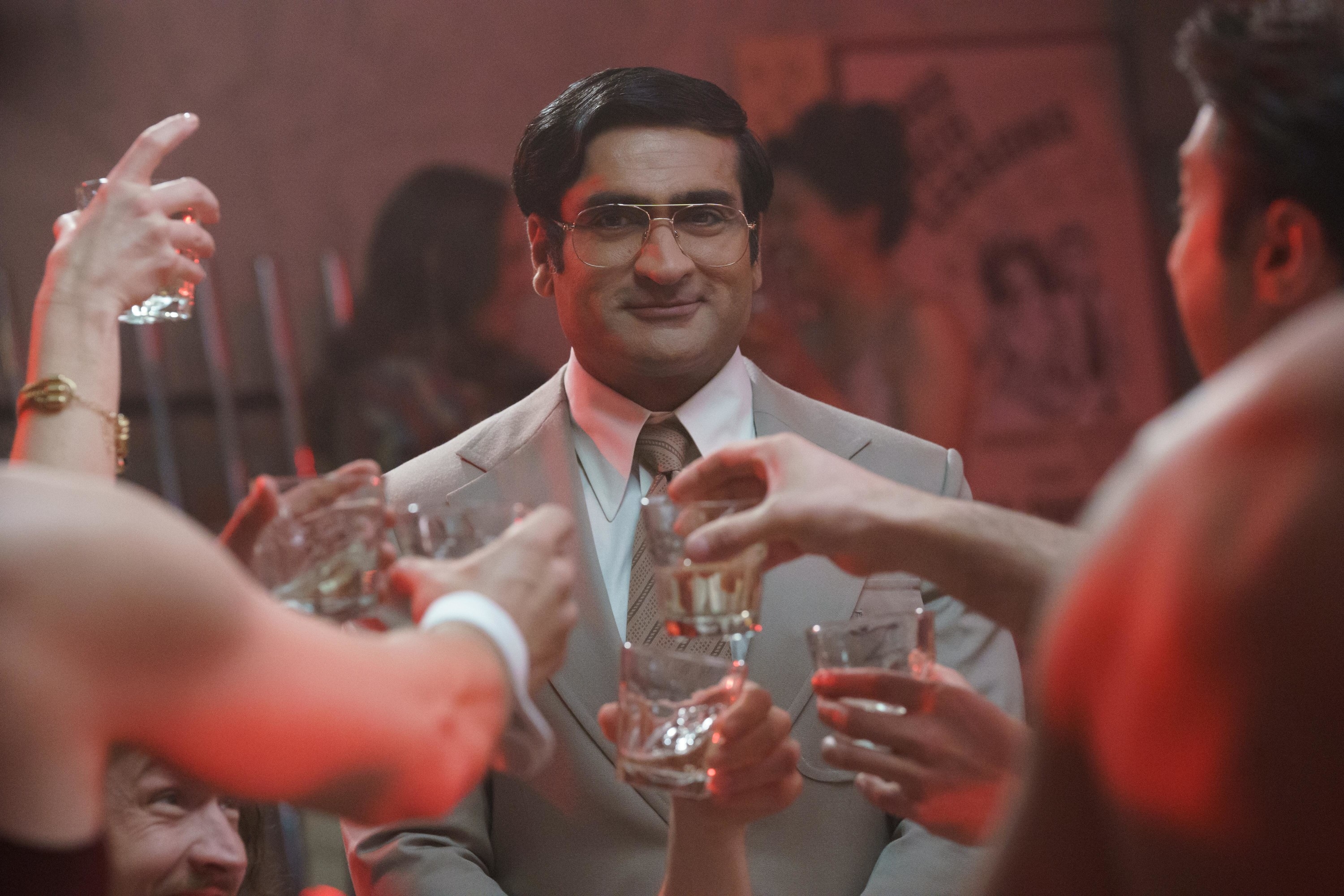 Kumail Nanjiani smiling in a bar as people clink their glasses