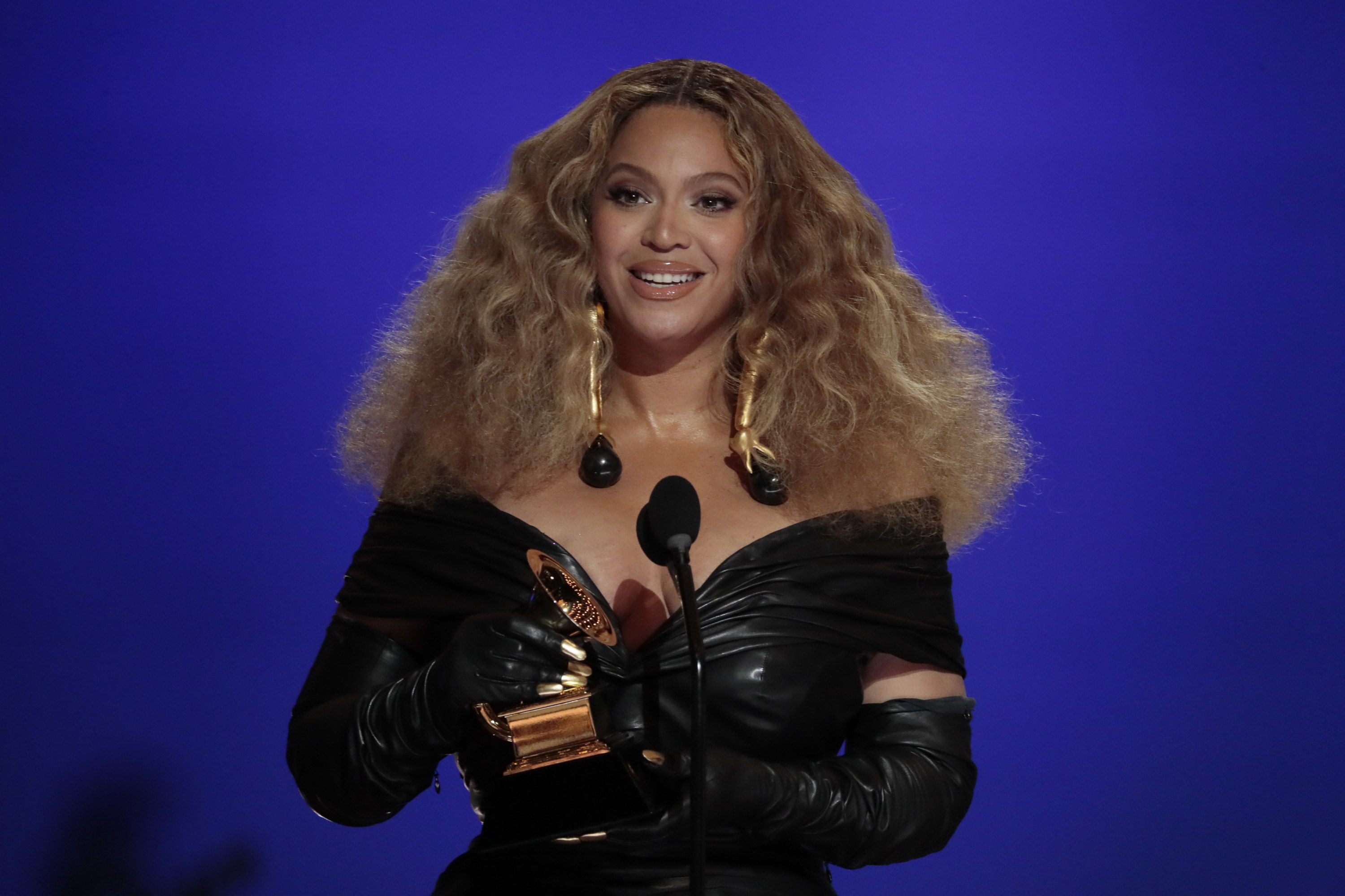 Beyonce wears a black latex off the shoulder V-neck dress with long sleeves. She is smiling mid-speech as she accepts an award. Her dark blonde hair is full and wavy, falling to her shoulders.