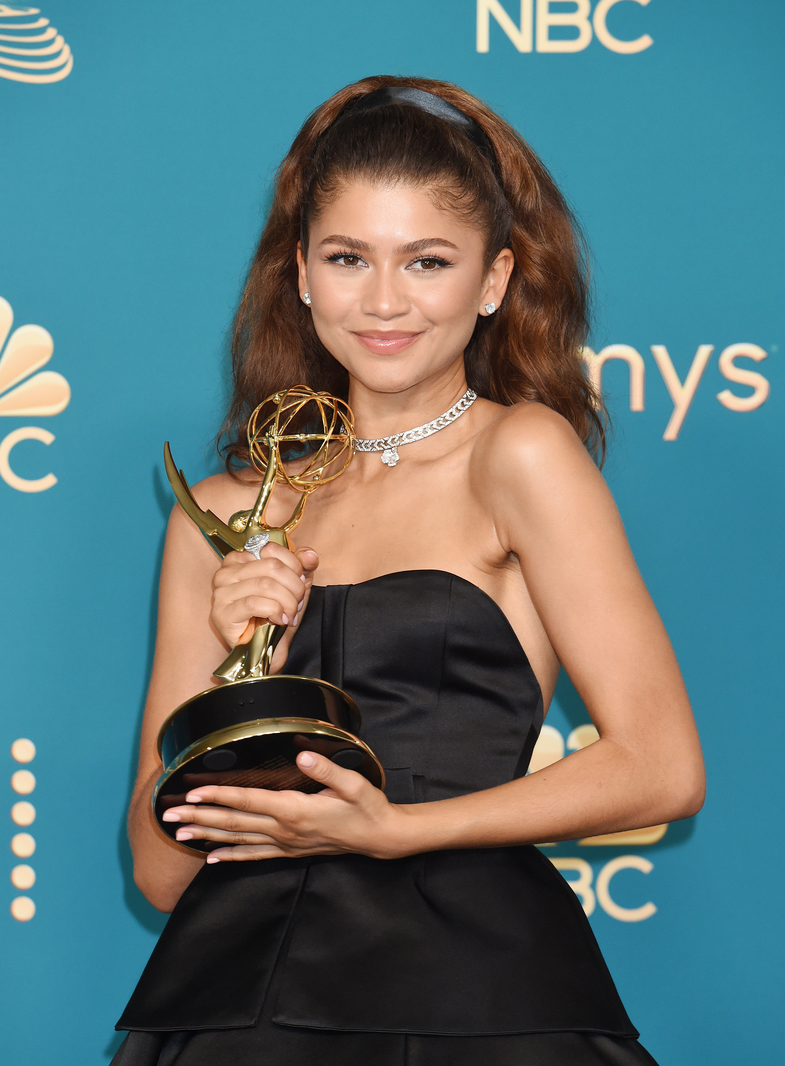Zendaya wears a black dress fitted in the torso and flowing outwards from the rib cage. She holds an award and smiles, her medium brown hair pushed back from her face by a black ribbon. She wears a chunky silver jewellery,