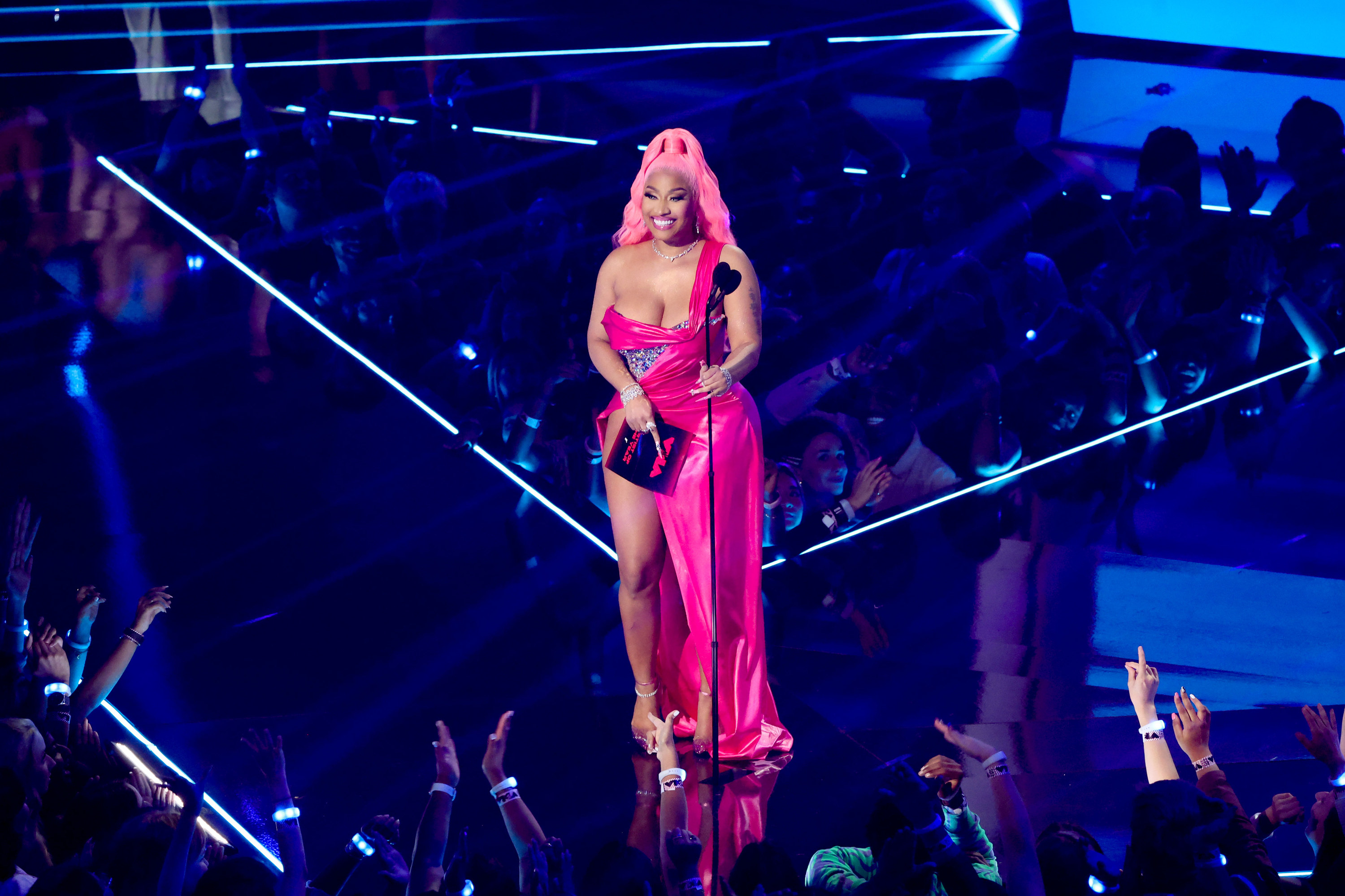 Nicki stands in the middle of a darkened stage with the spotlight illuminating her. She wears a bright pink cocktail dress with one leg showing, and her hair is candy pink and flows down her back from a tall ponytail atop her head. She is smiling widely.