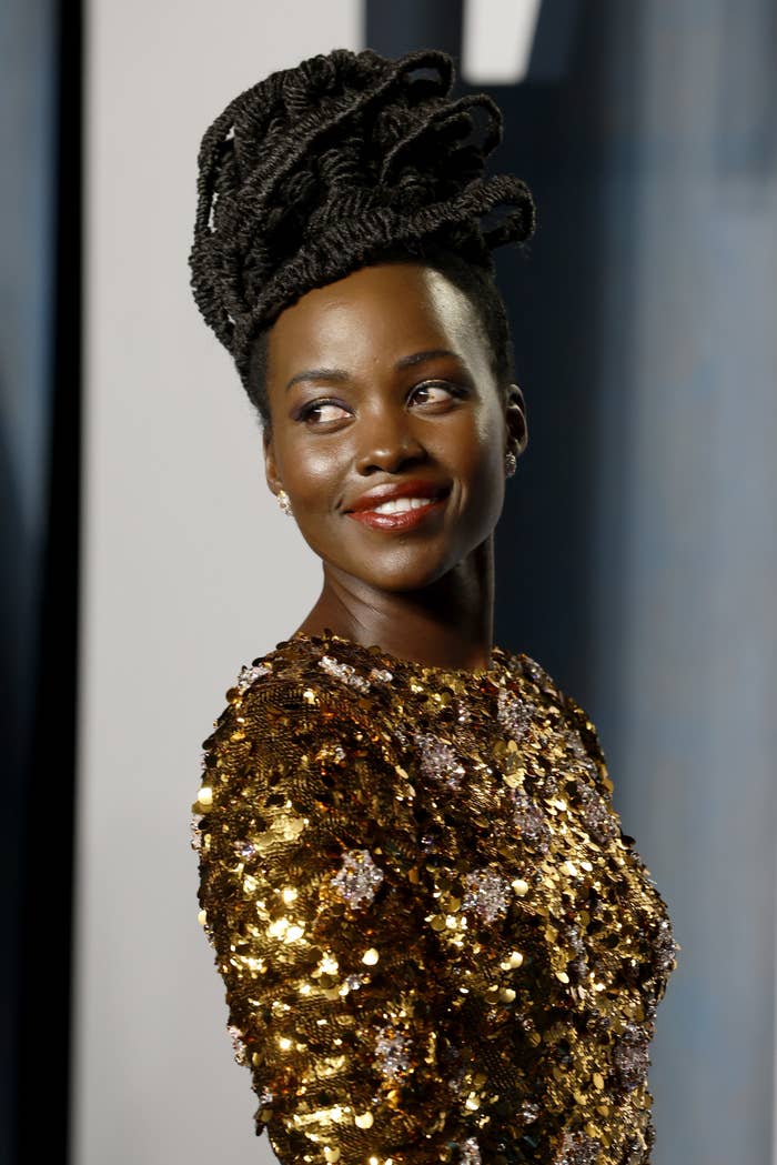 Lupita wears a gold sequinned long sleeved dress. She is side on, looking back over her shoulder and smiling. She wears a deep red lipstick and her hair is in an intricate braided up do.