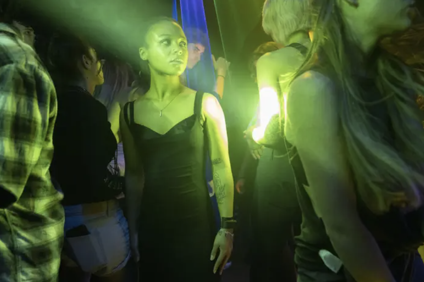 a woman wearing a black dress stands in a crowded club bathed in green light