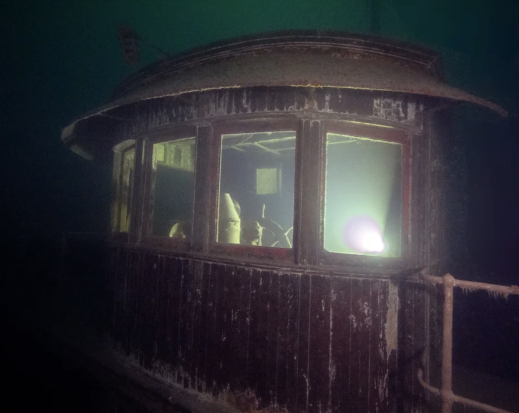 A helmsman&#x27;s cockpit of a ship with a huge steering wheel; a light is also reflecting off a window and makes it seem like there&#x27;s a light on inside the cockpit