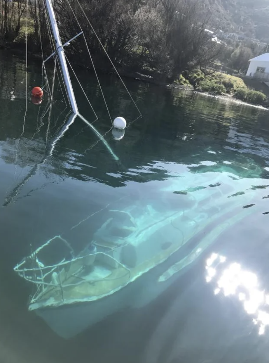 A boat with its mast visible above the water, but the rest of it is submerged and can be seen at the bottom of the lake