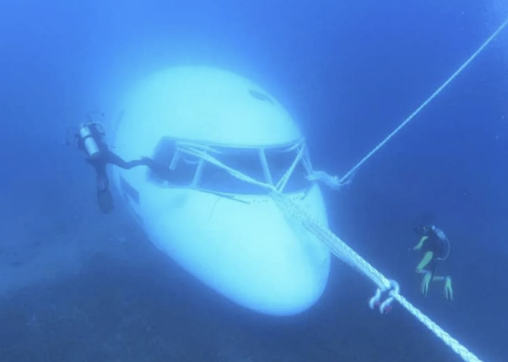 Scuba divers swim around a different submerged plane, with what looks like ropes attached to the front to pull it out of the water