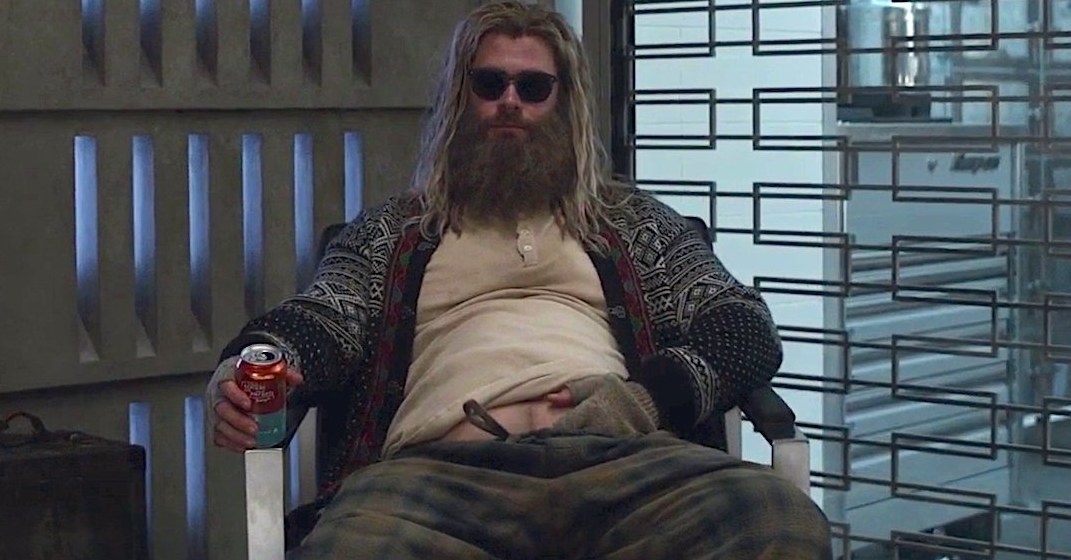 Fat Thor wears dirty clothes and sits with his hand tucked into his pants and sunglasses on with a soft drink in his hand