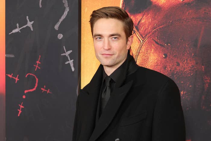 Robert wears a black coat over a black shirt and silky tie, and smiles in front of a black, yellow and red backdrop advertising his film "The Batman"