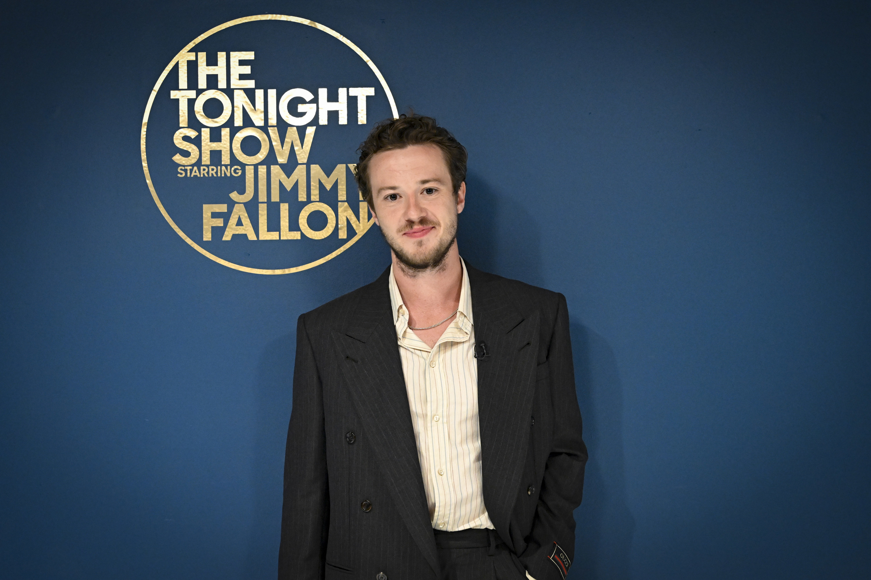 Joseph wears an oversized black blazer with a yellow and white pinstripe shirt; he smiles, standing in front of a navy wall with a gold sign reading &quot;The Tonight Show starring Jimmy Fallon&quot; in a circle