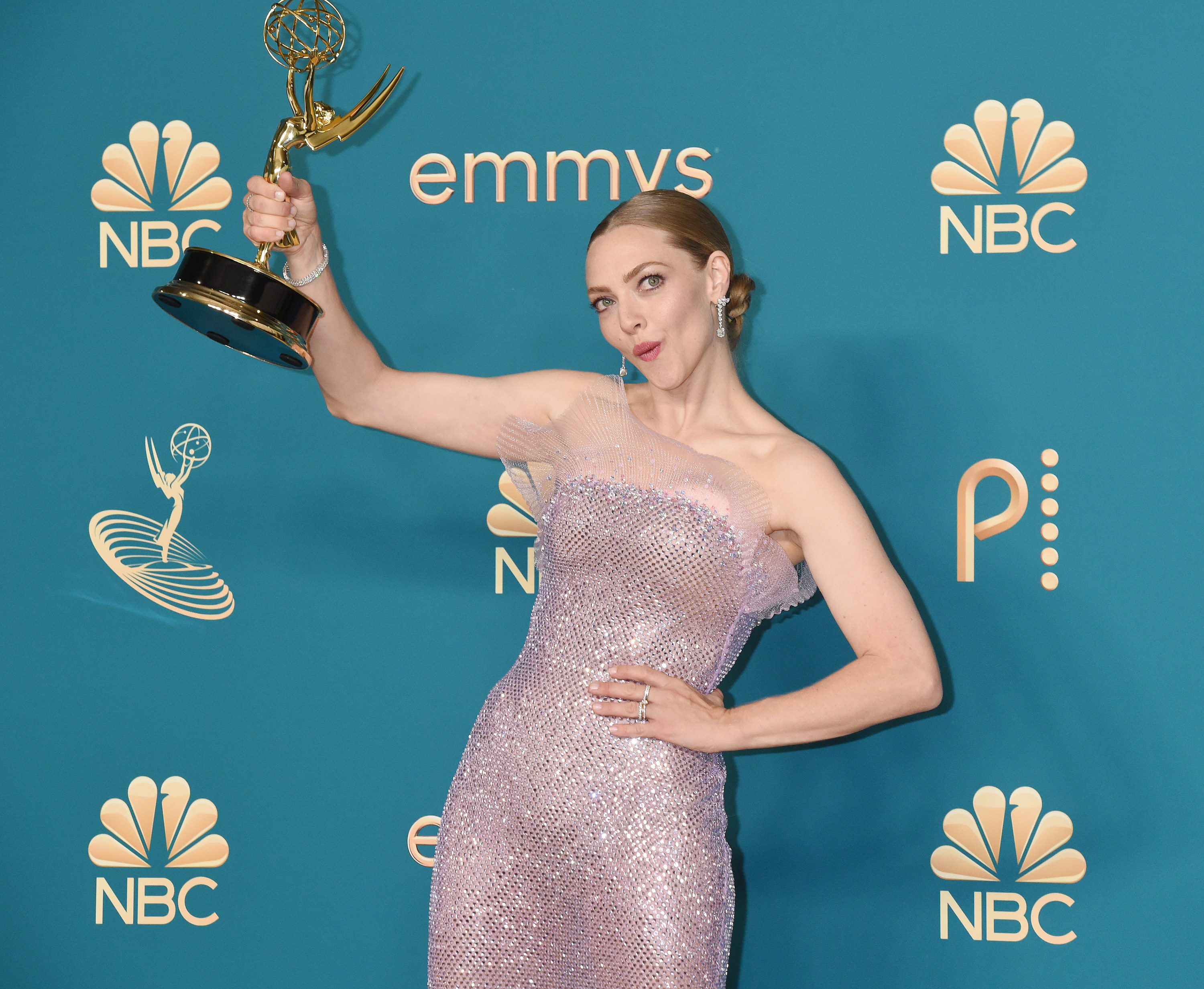 Amanda wearing a glittering sleeveless bodycon dress in a pale lilac color, holding her Emmy victoriously; her blonde hair is tied back in a bun and she wears silver drop earrings