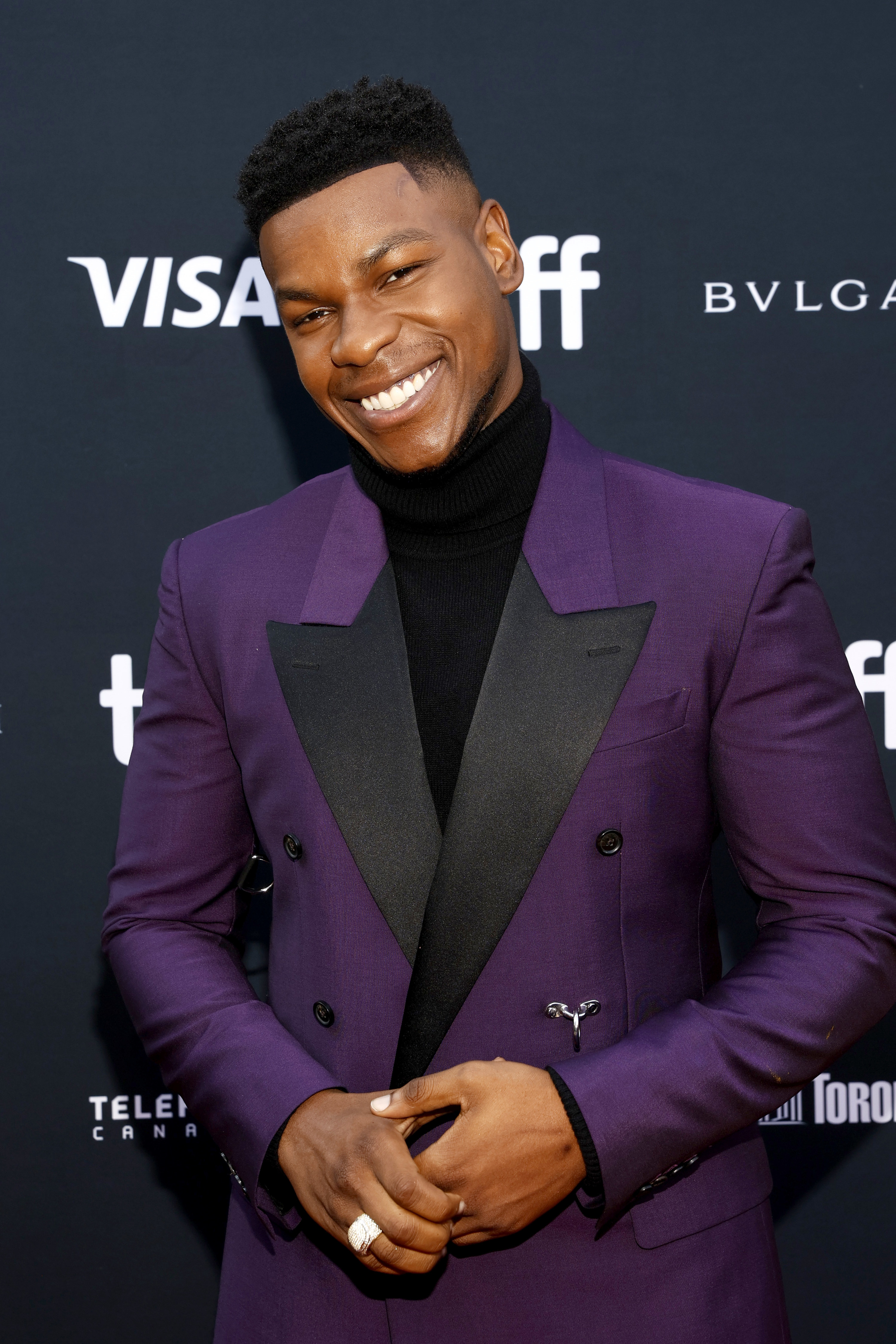 John smiling broadly, his hands clasped together at his waist; he wears a purple suit with black lapels and a black turtleneck underneath, with a chunky silver ring on one finger
