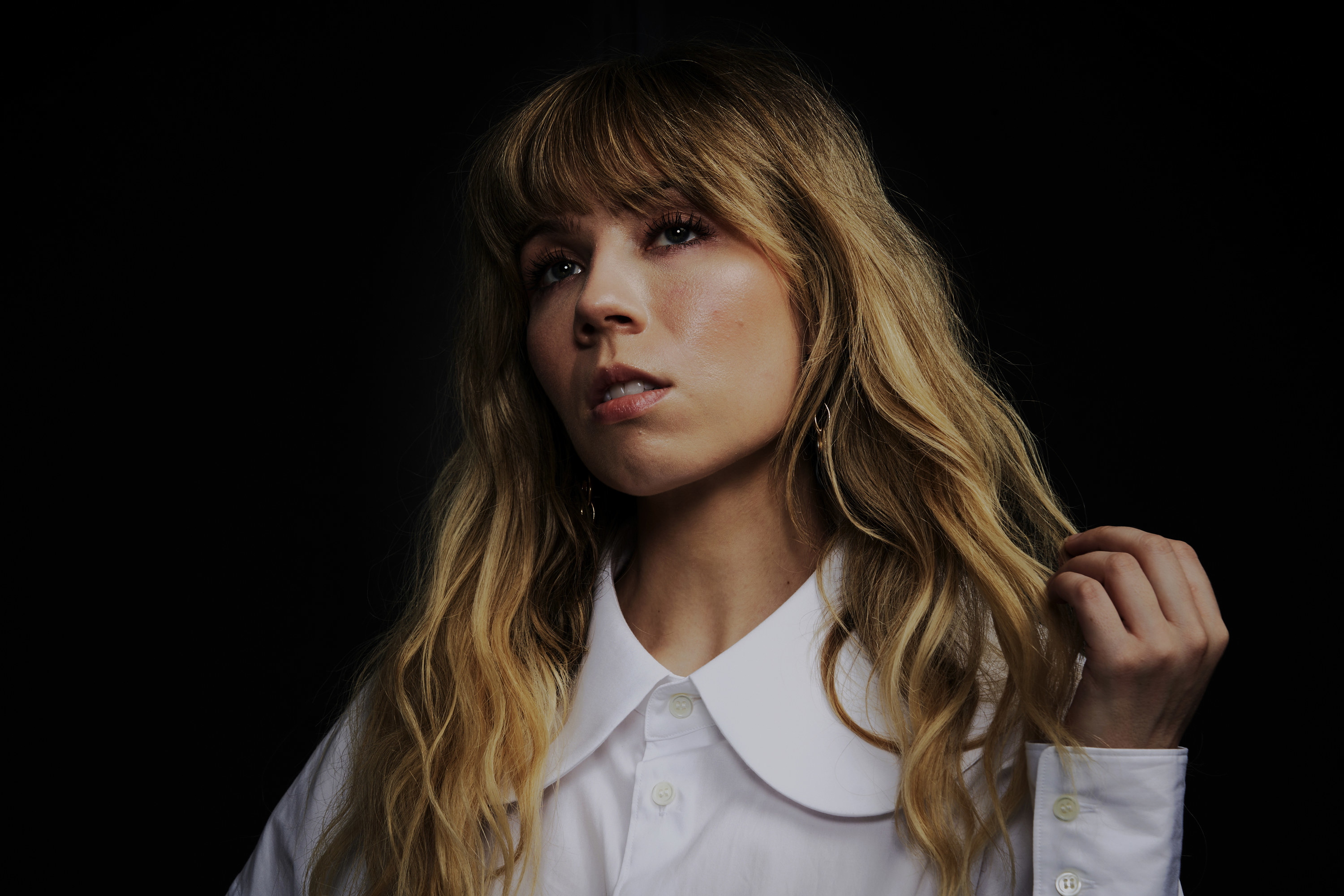 Jennette poses in front of a black background, looking into the distance with a serious expression; her mouth is slightly open and her long blonde hair is wavy, falling past her shoulders onto the white blouse she wears.