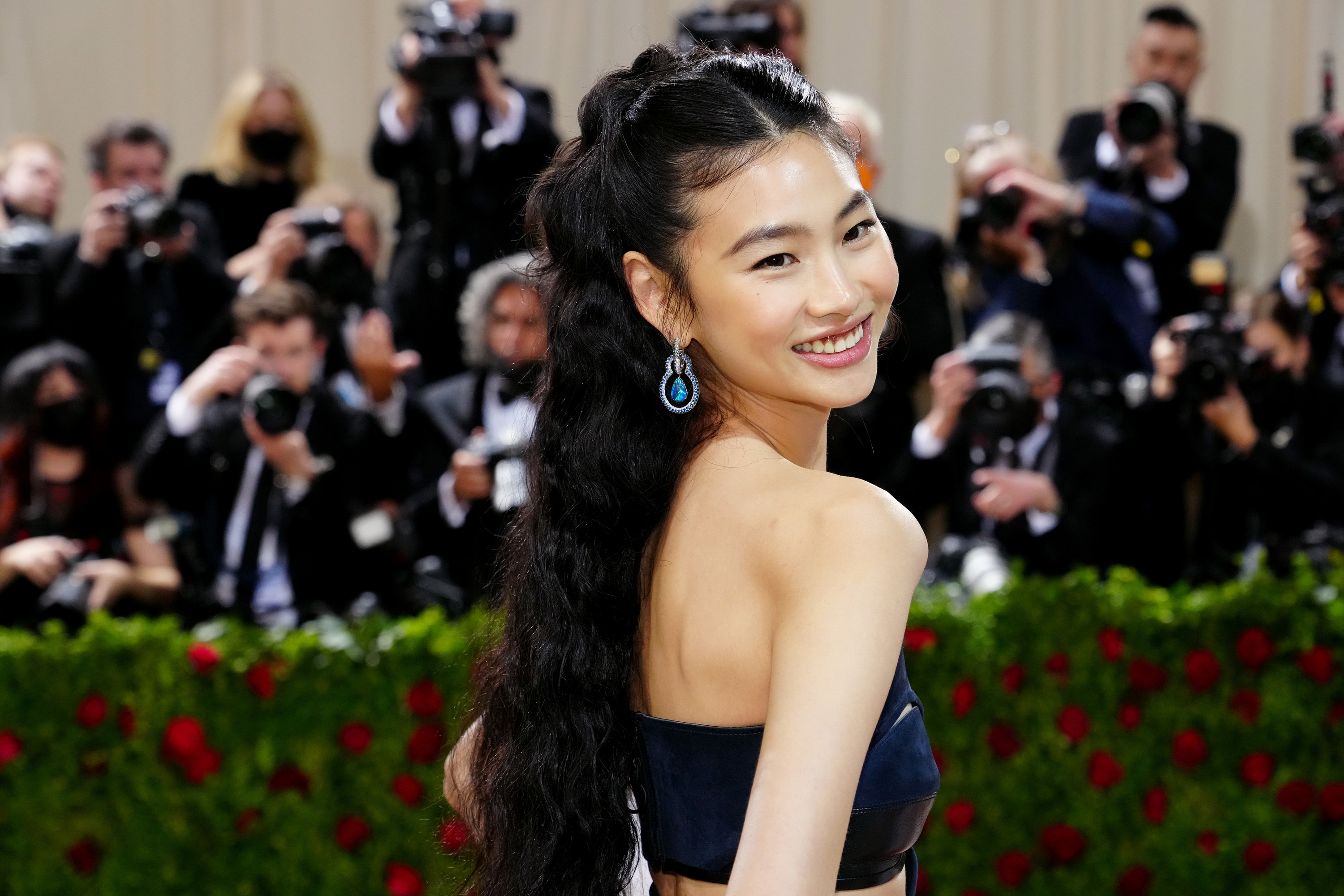 Hoyeon looks back over her shoulder, her long dark hair tied up and falling in glossy waves down her back; she is wearing a navy tube dress with blue and silver drop earrings; she is smiling broadly and has her hands on her hips