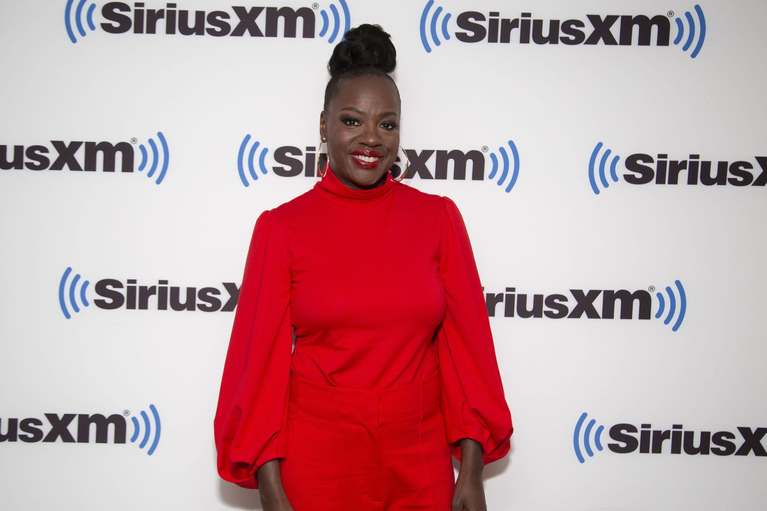 Viola wears a red high-neck, balloon sleeved blouse with red trousers; her hair is in a high bun atop her hair and she wears red lipstick; she is smiling and stands in front of a wall with Sirius XM on it
