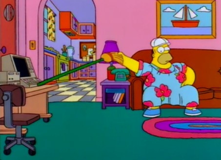 Homer wears a muumuu and uses a broom to tap his computer while seated on the couch