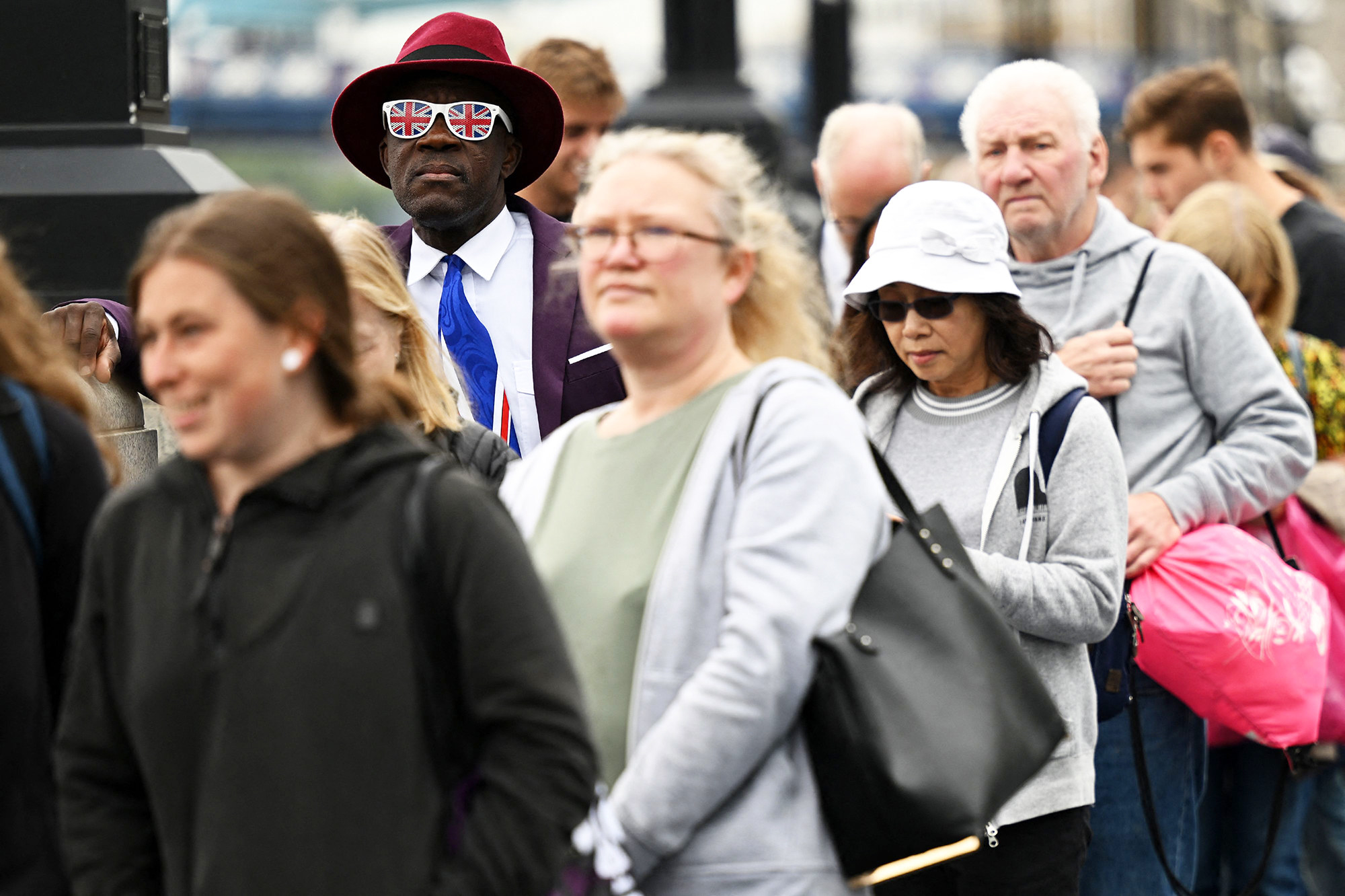 Man wearing a hat and Union Jack sunglasses waits in a crowd