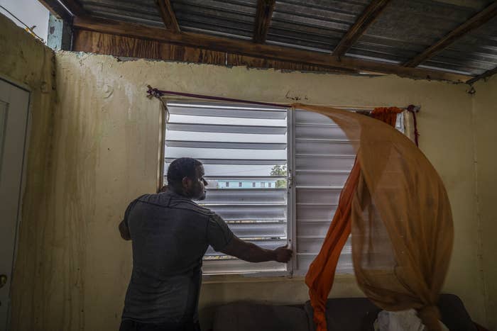 A man stands at a small window with horizontal blinds and blowing curtains