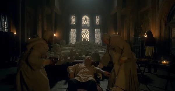 Viserys reclines on a chair looking ill while two Maesters stand over him