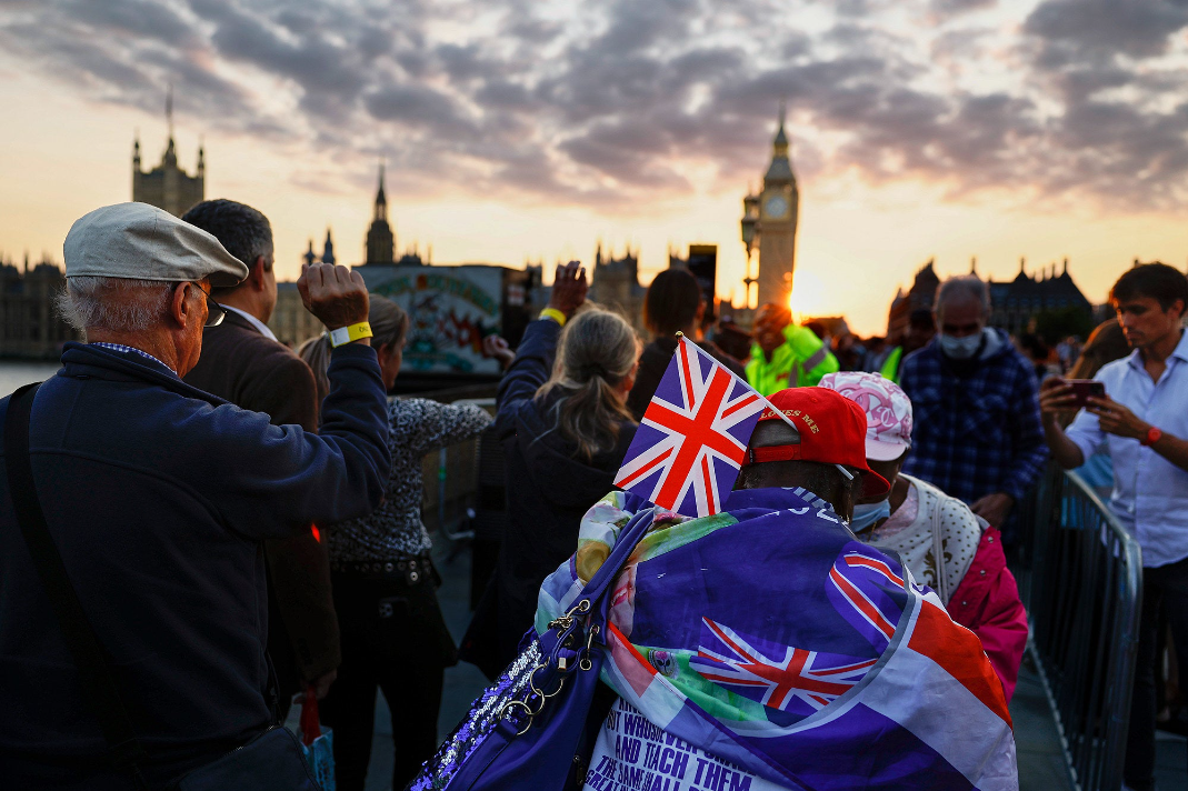 A crowd looks toward Big Ben at sunset, one person is kitted out in Union Jack gear