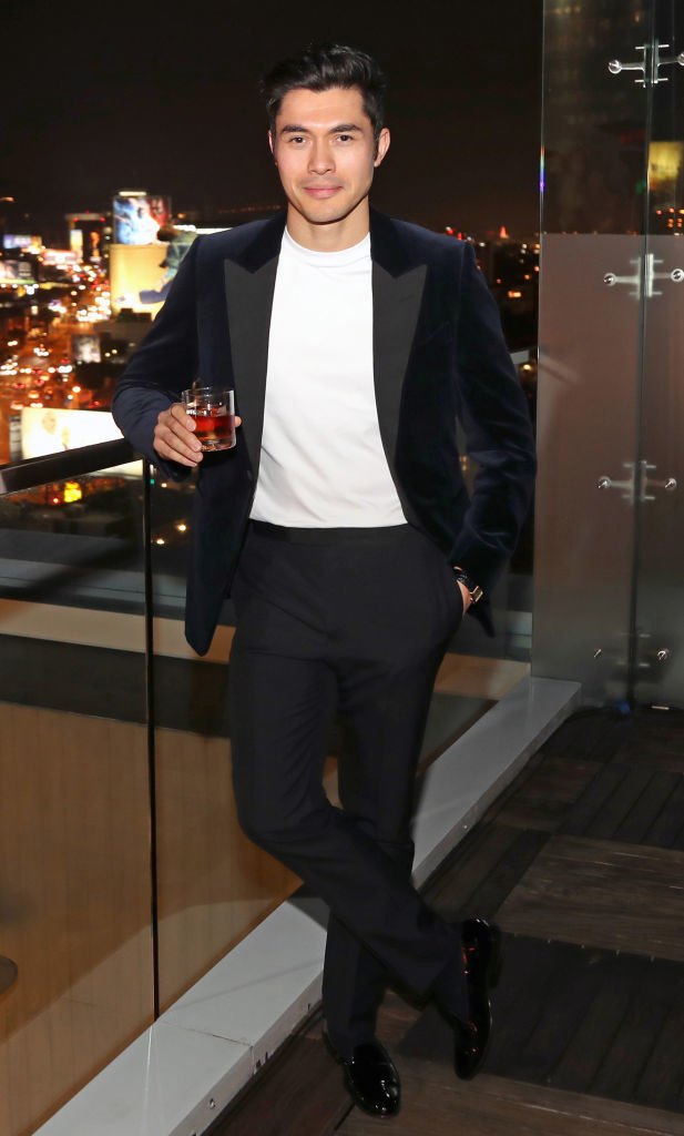 Henry in a suit jacket and T-shirt and holding a drink in front of a city scene at night