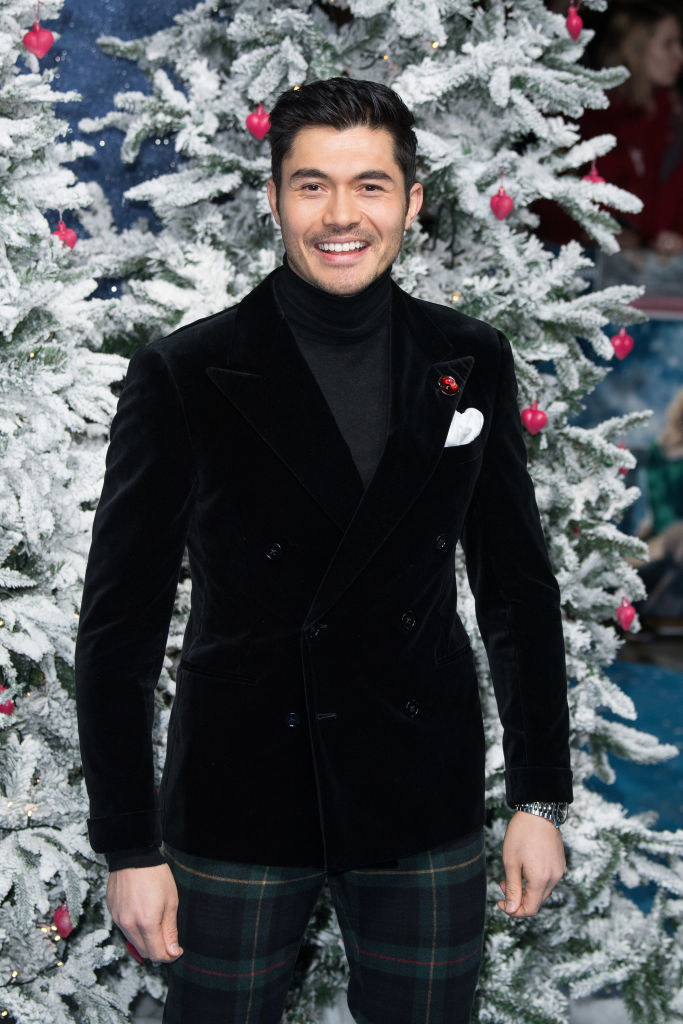 Henry smiling and wearing a velvet jacket and plaid pants in front of a large Christmas tree