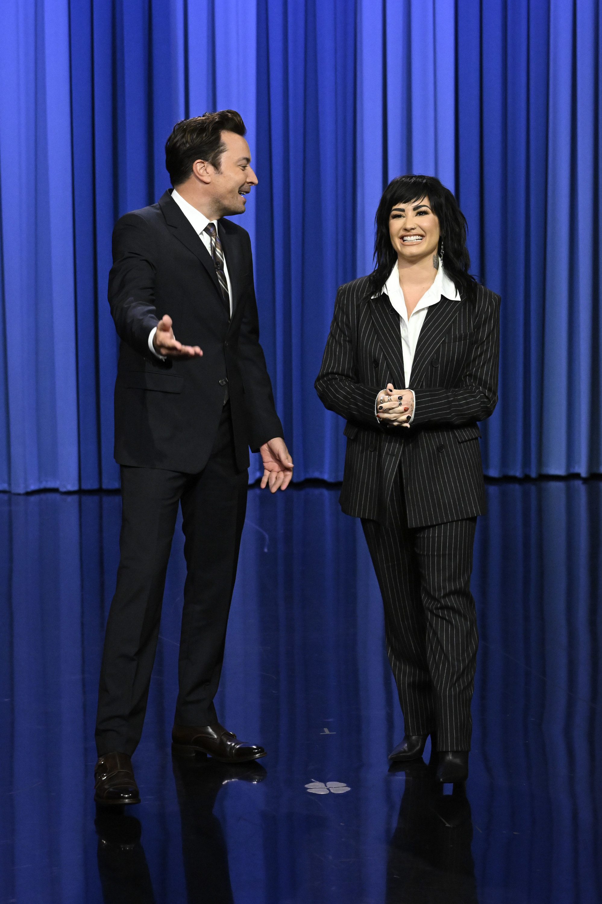 Demi wearing a striped pantsuit and smiling with Jimmy Fallon