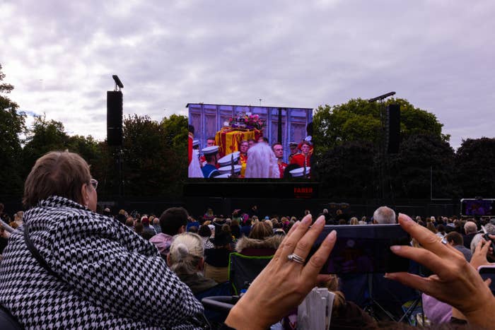 An audience sits on a lawn watching a big screen on which the queen's funeral plays
