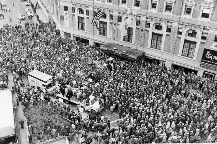 A street is crowded with people in front of a hotel in this aerial black-and-white image