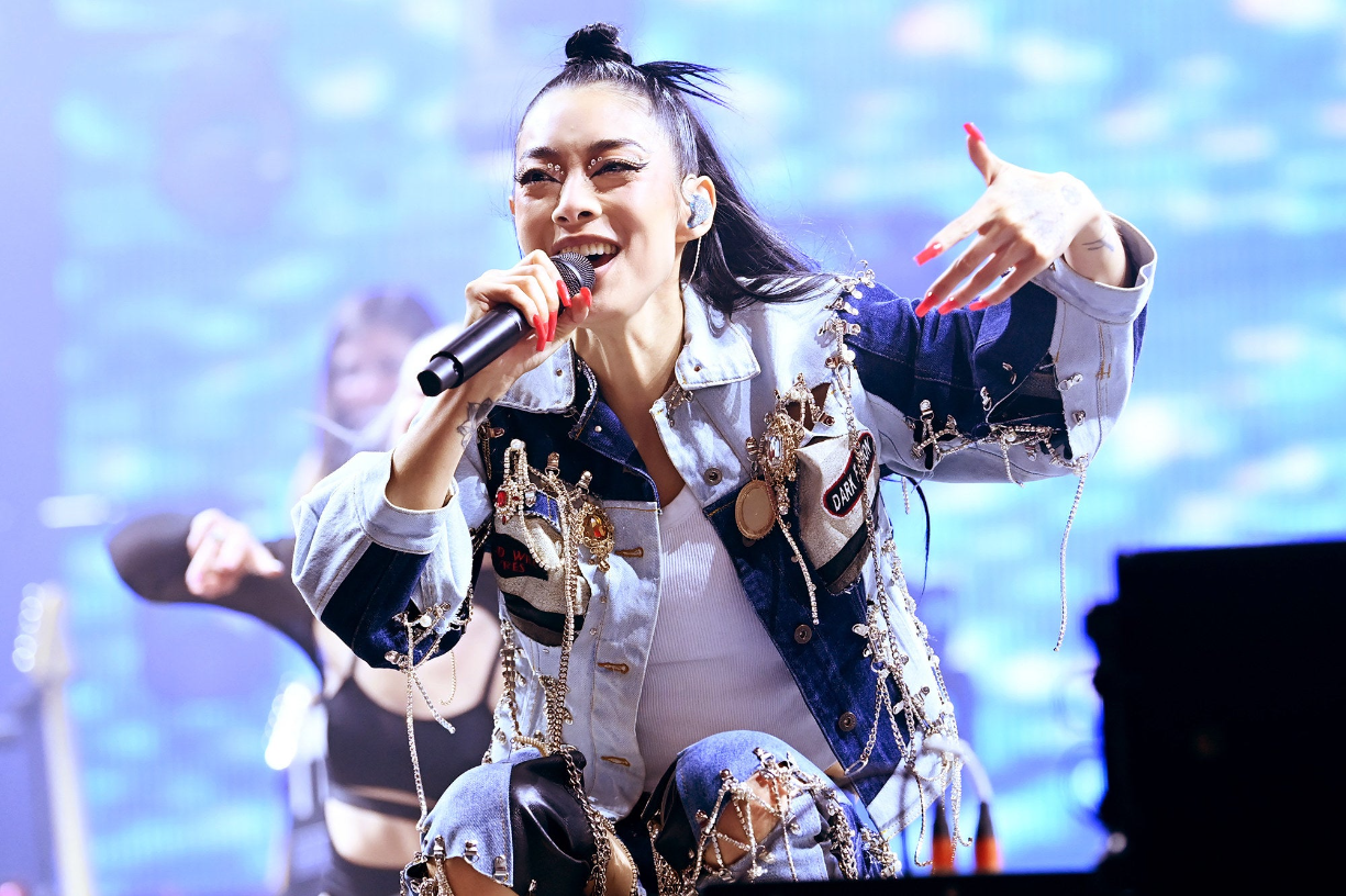 Rina wears an incredible denim and leather jacket and pants combo while crouching onstage and singing into a mic