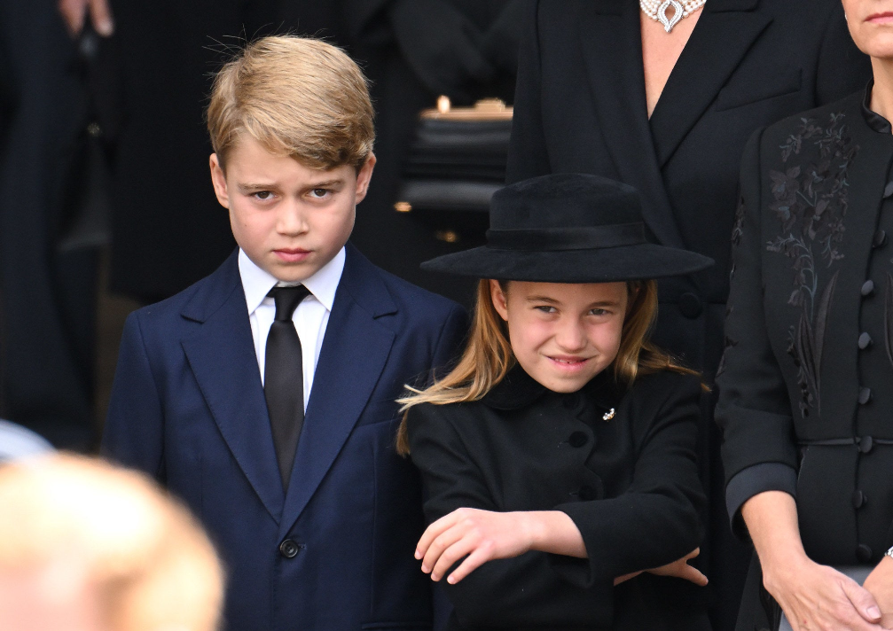 young royal boy in a navy suit looking pensive, next to him is a grinning young girl in a black outfit and accompanying hat