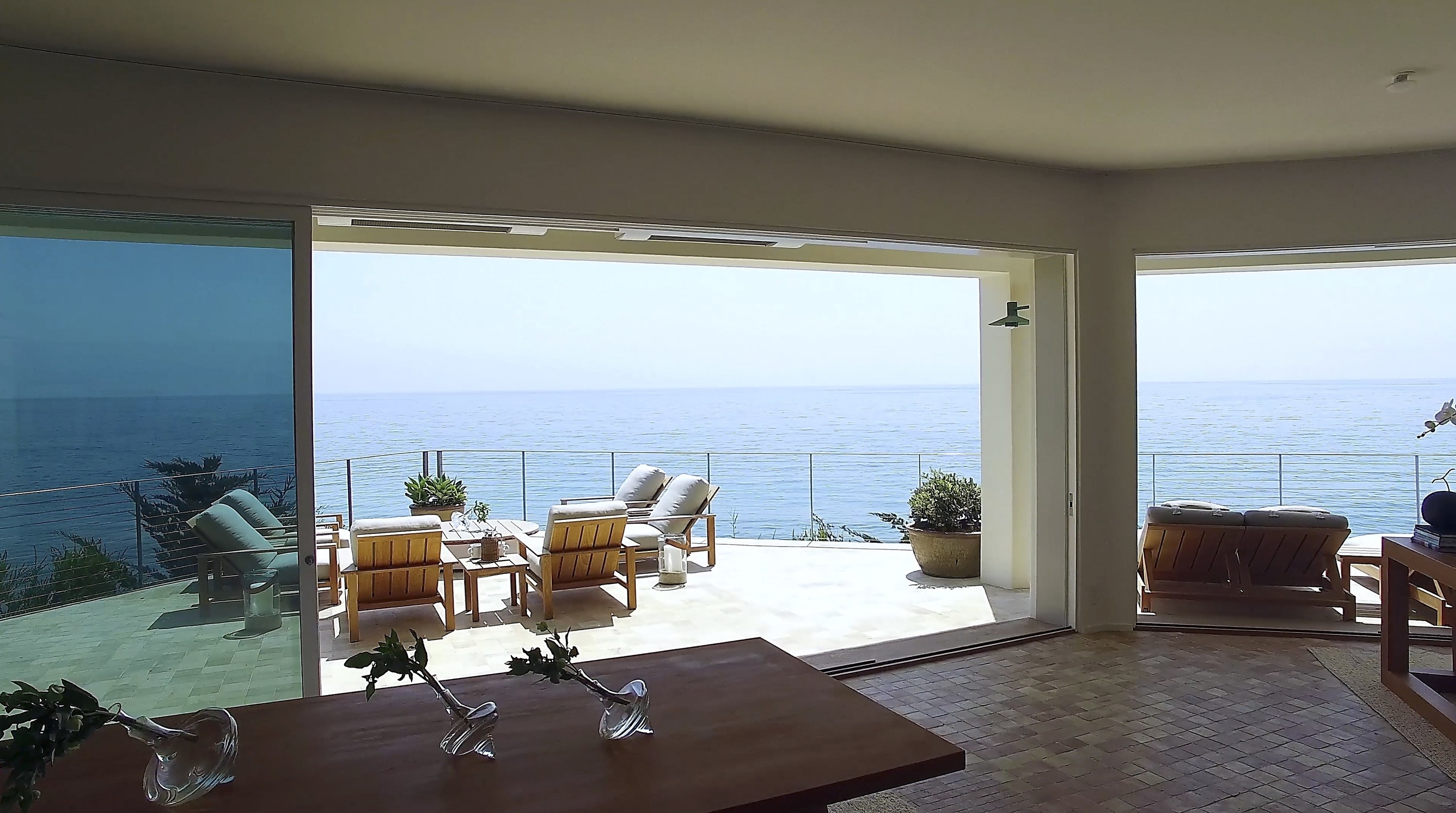 A view from inside the house shows a patio area with table and chairs overlooking the ocean