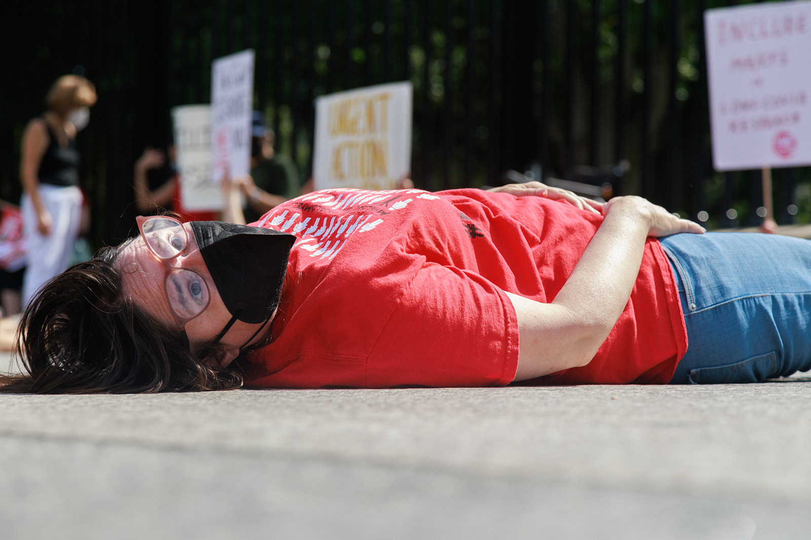 A person in a red shirt lying on the ground with a black mask on during a protest about COVID-19