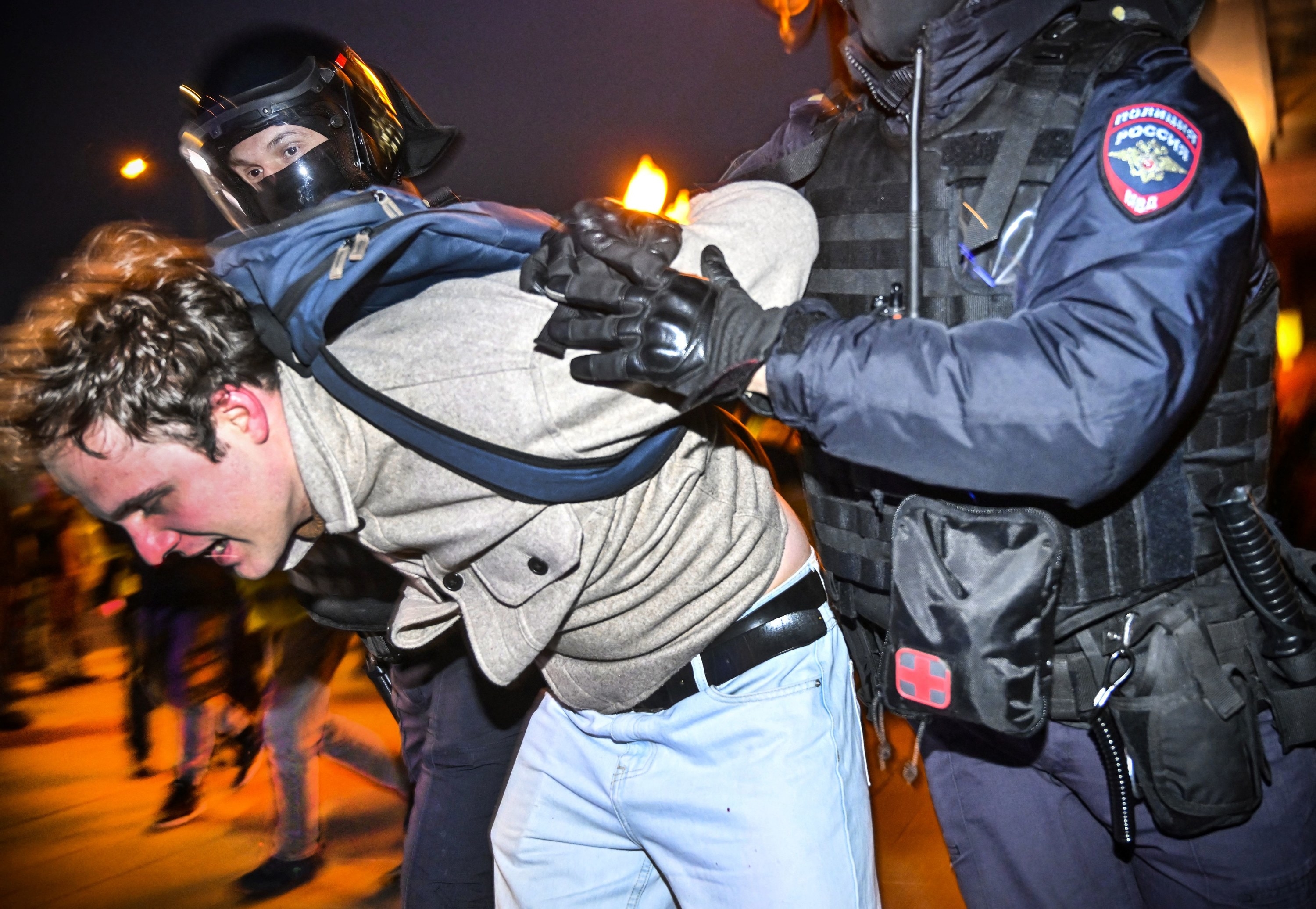 A police officer in SWAT garb restrains a man in an oatmeal sweater who is doubled over at a protest