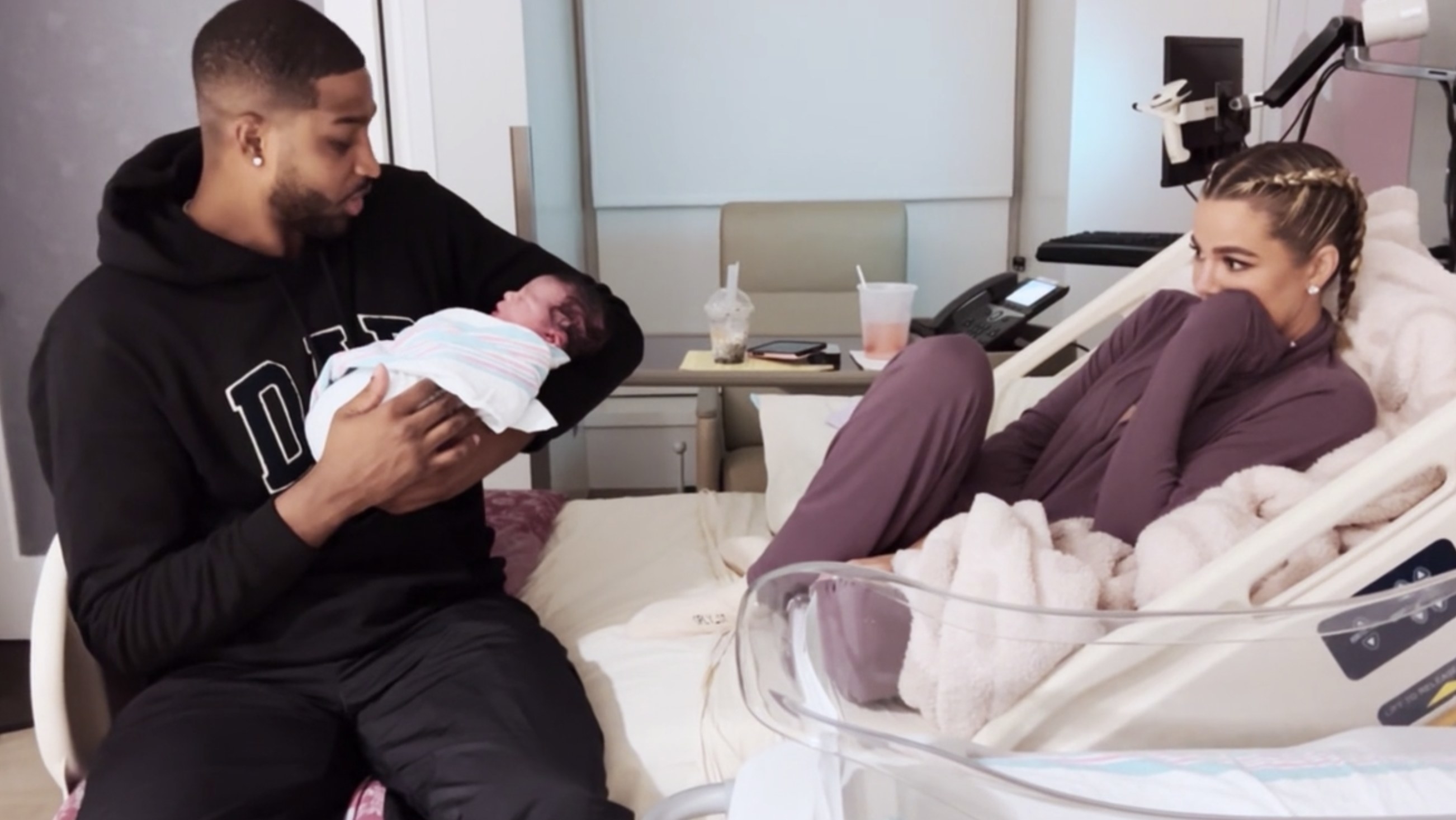 Tristan sits on the edge of the bed holding the baby while Khloe looks on