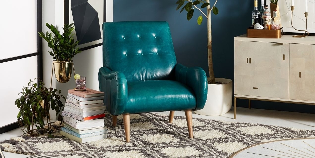 tufted dark blue chair with wood legs in corner of room next to stack of books