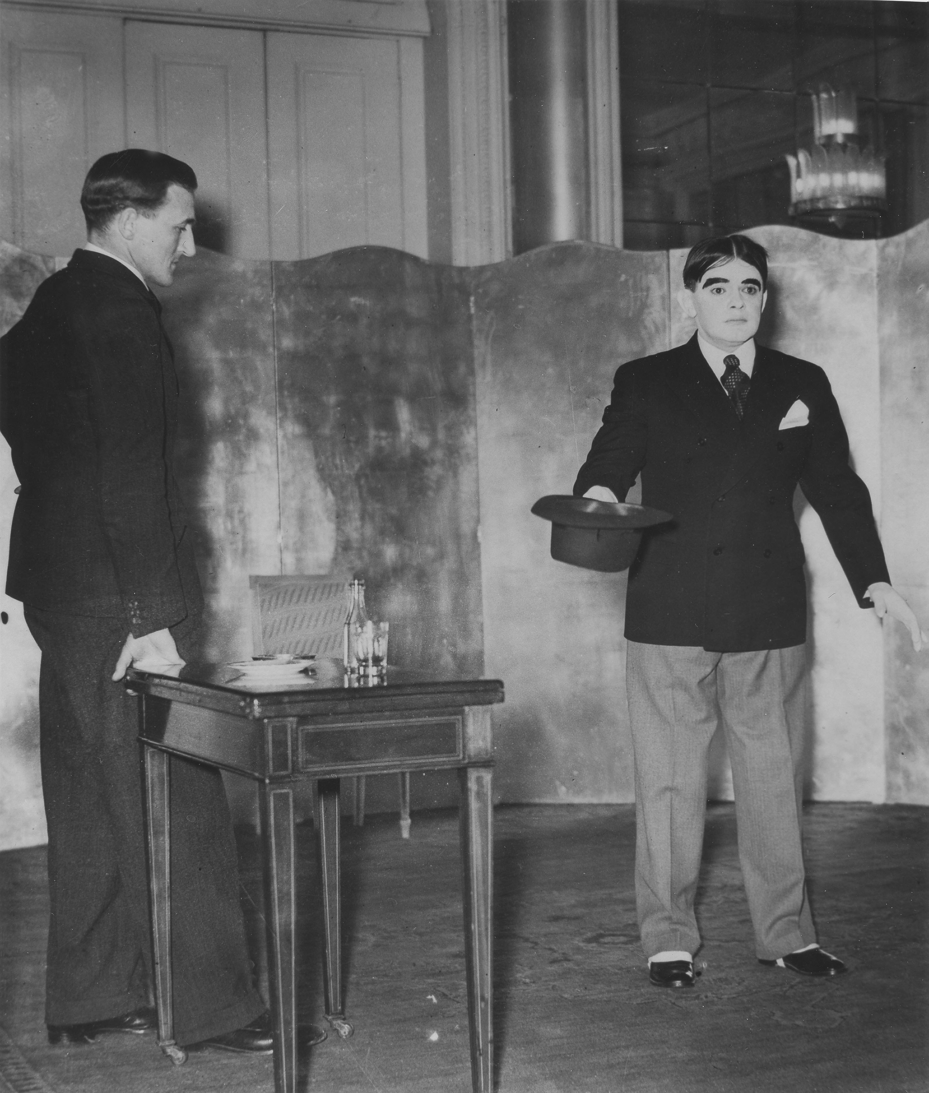 A black-and-white photo of a man in a suit standing at a small table, looking at another man who stands with stiff posture and limbs and a blank expression, his face painted white