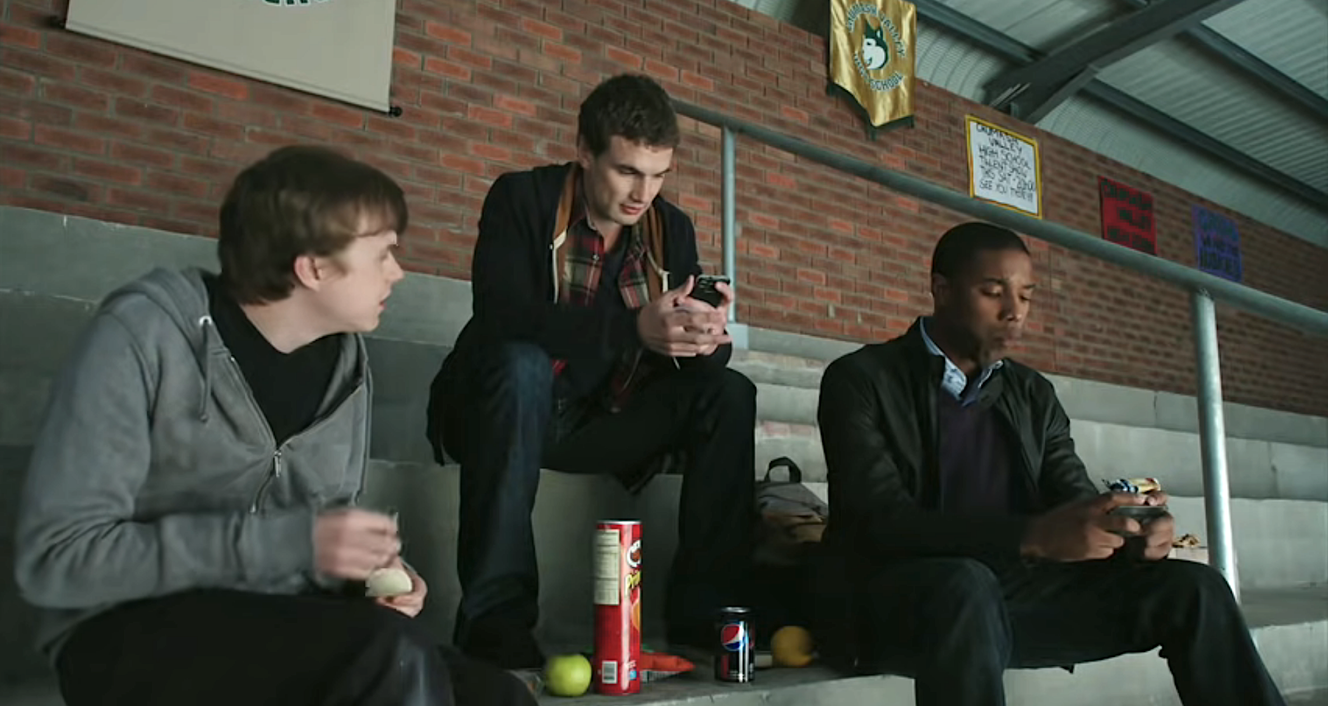 Three young guys sitting in the bleachers of a gym eating and talking and looking at their phones