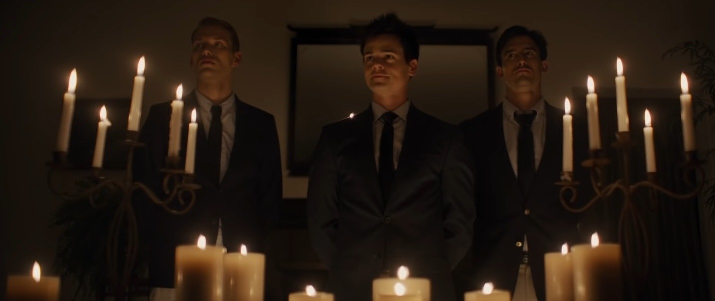 Three scary-looking fraternity brothers stand in full suits in front of a candlelit room in &quot;Pledge&quot;