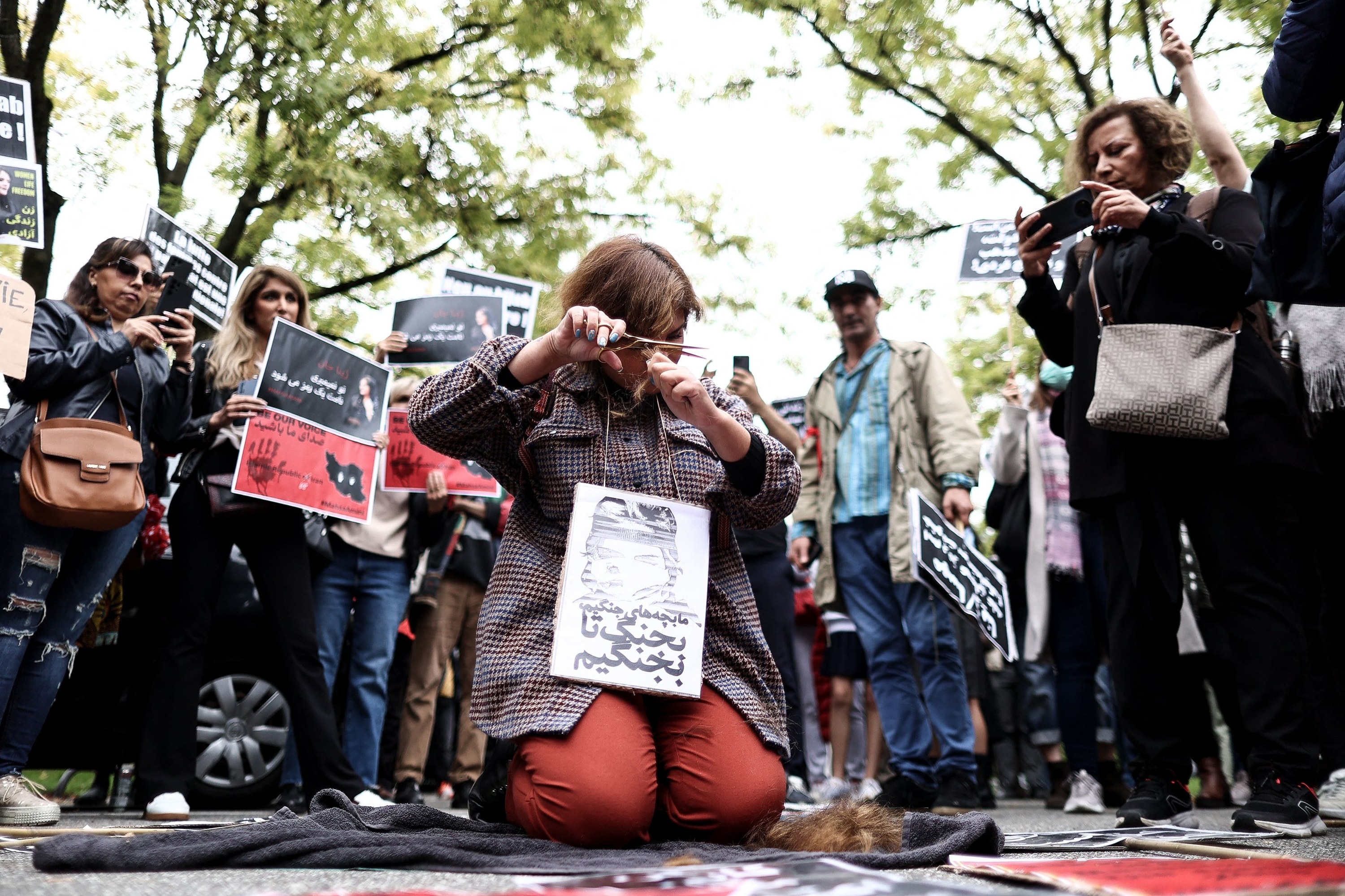 A woman kneels on the ground and cuts her hair in front of a crowd of protesters