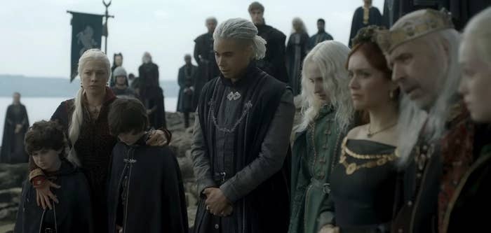 Laenor, Rhaenyra, Luke, Jace, Aegon, Alicent, Viserys and Helaena stand in a row in front of a crowd of people, all looking sad