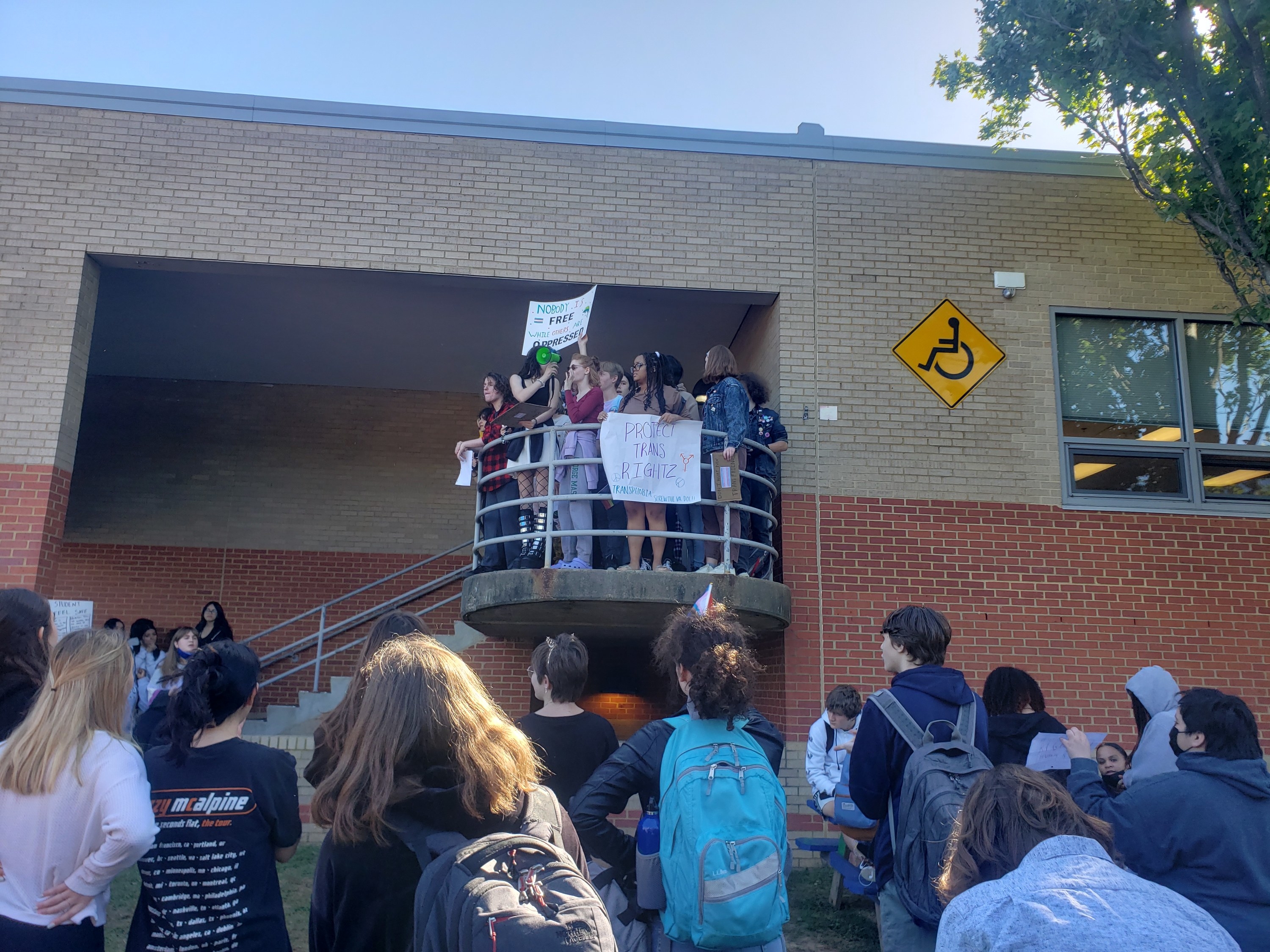 Students crowd outside a school, listening to peers speaking from a platform