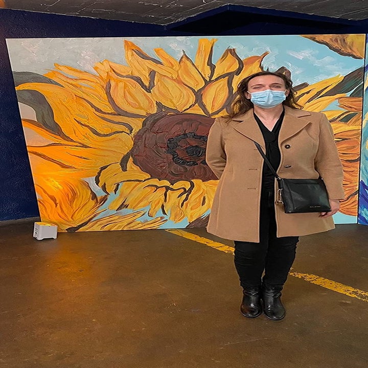 A different reviewer wearing the tan coat and standing in front of sunflower painting